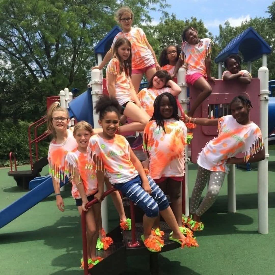 A group of people wearing matching orange tie-dye shirts pose together on a playground structure on a sunny day.