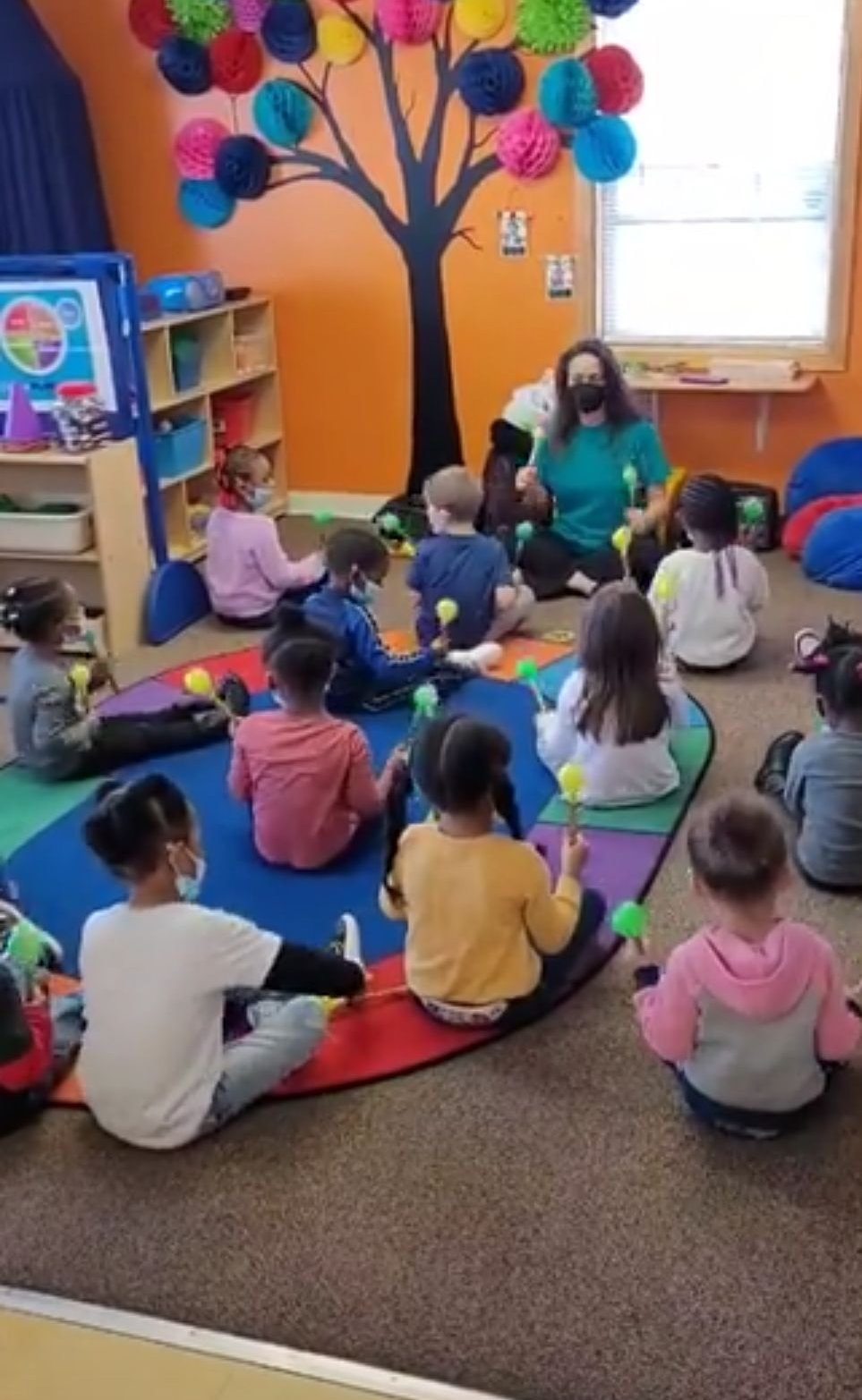 A teacher and a group of children sit on a colorful rug in a classroom, each holding small yellow percussion instruments.