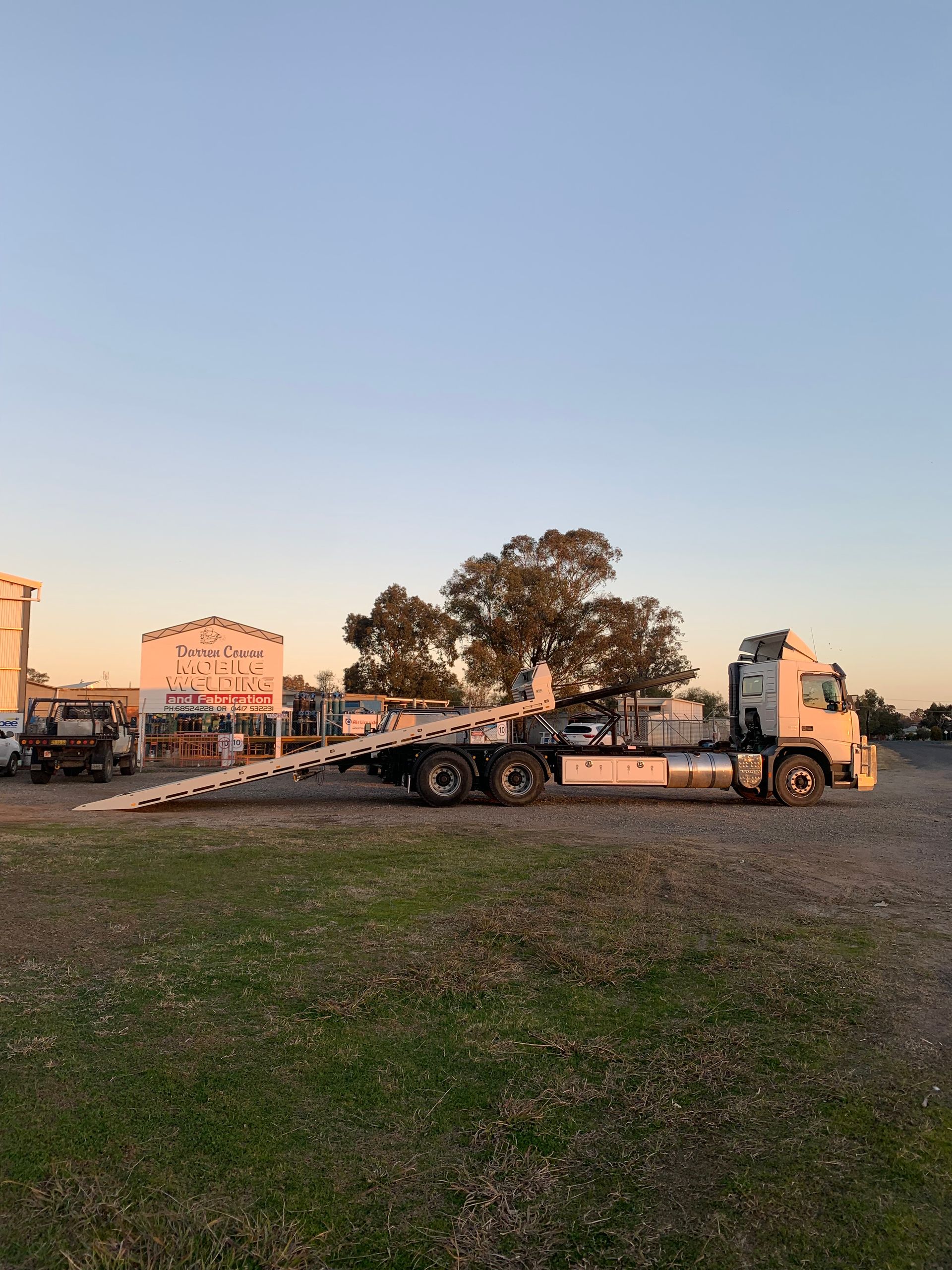A flatbed tow truck parked on a grassy area with a building and trees in the background under a blue sky.