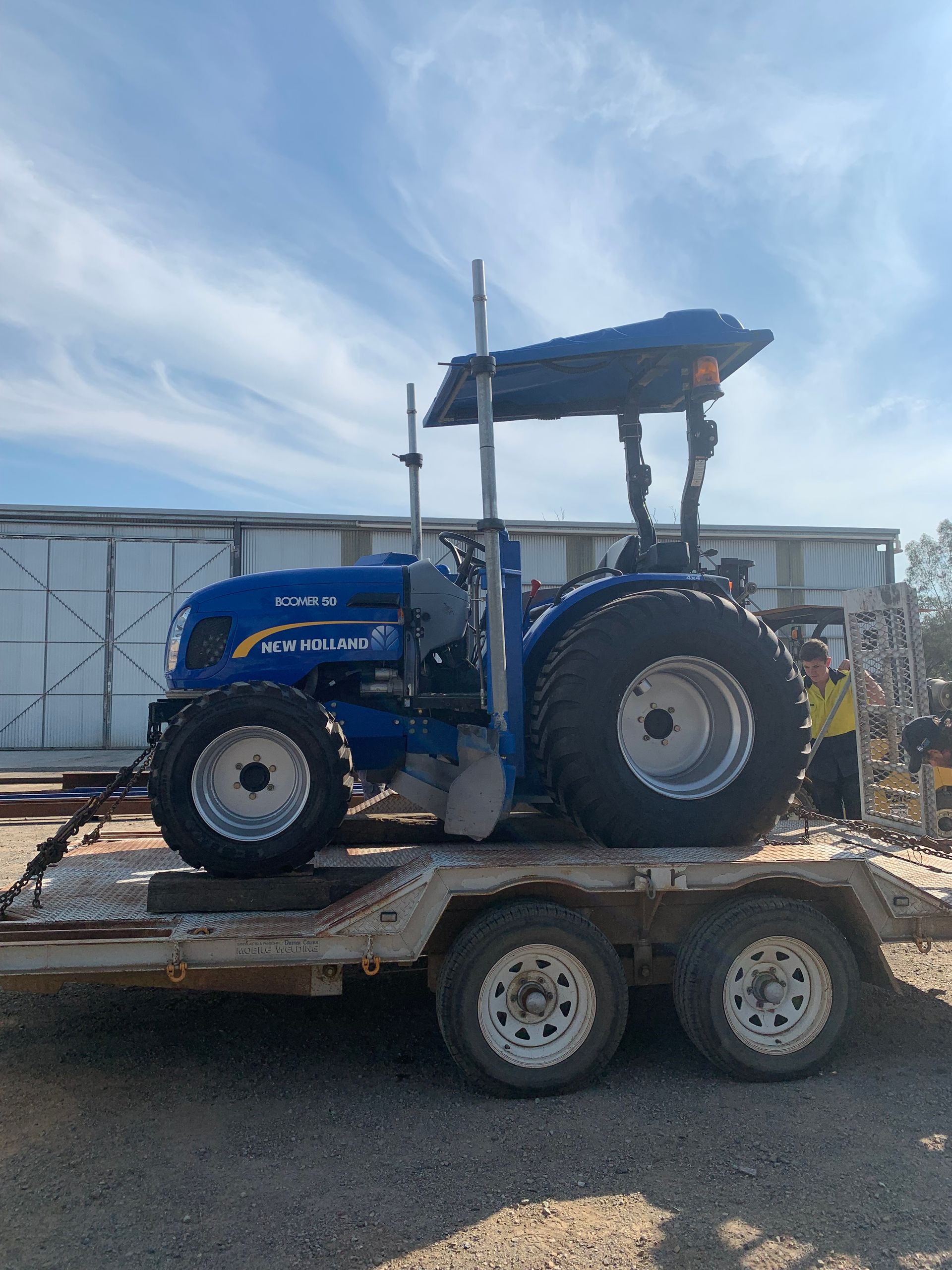 A blue tractor on a trailer, parked outside on a sunny day. A person is visible beside the tractor.
