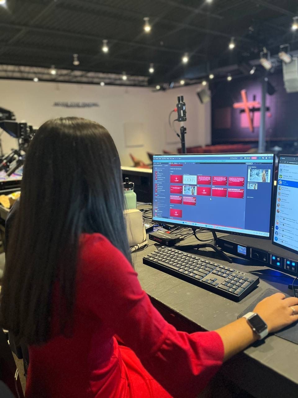 Woman in red dress operating computer in a studio, with monitors displaying graphics and a cross in the background.