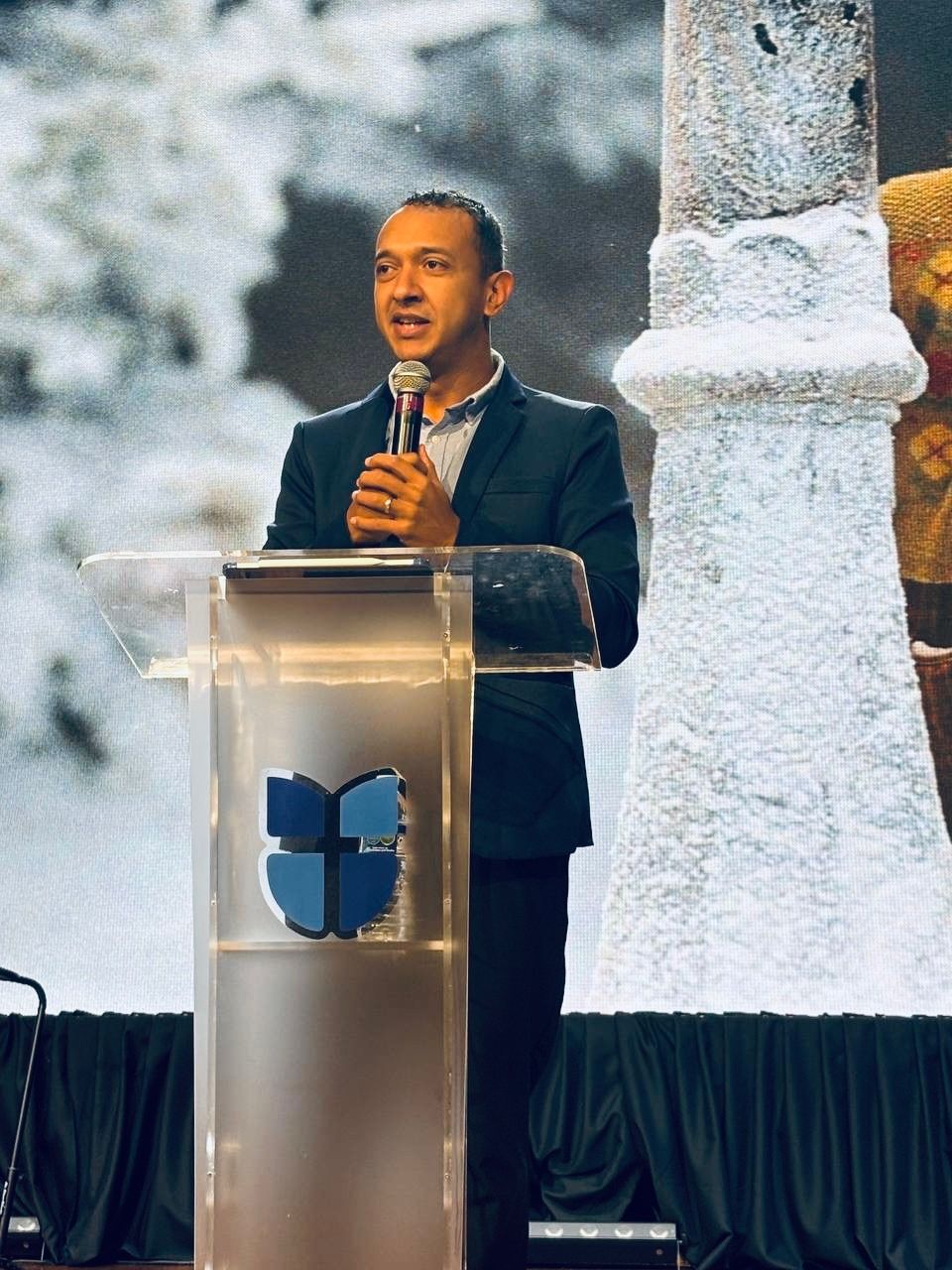 Man speaking at a podium with a logo on it, large screen with snow scene background.