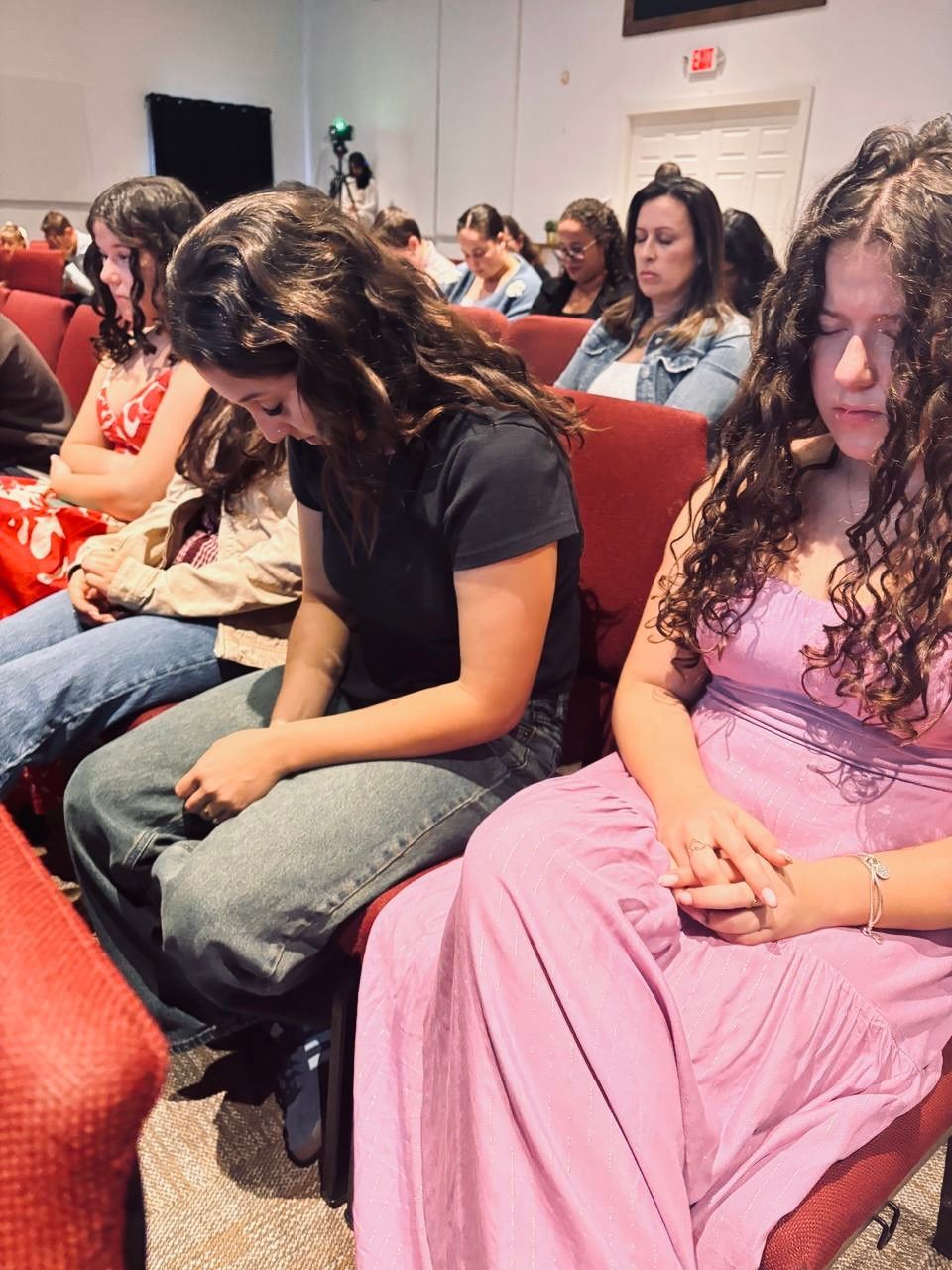 People with eyes closed, heads bowed, seated in a church. Red chairs, many more people in background.