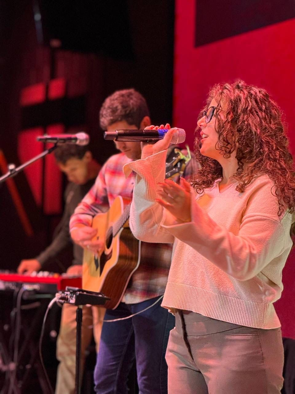 Band on stage: Singer with curly hair, guitarist, keyboardist. Red backdrop, stage lighting.