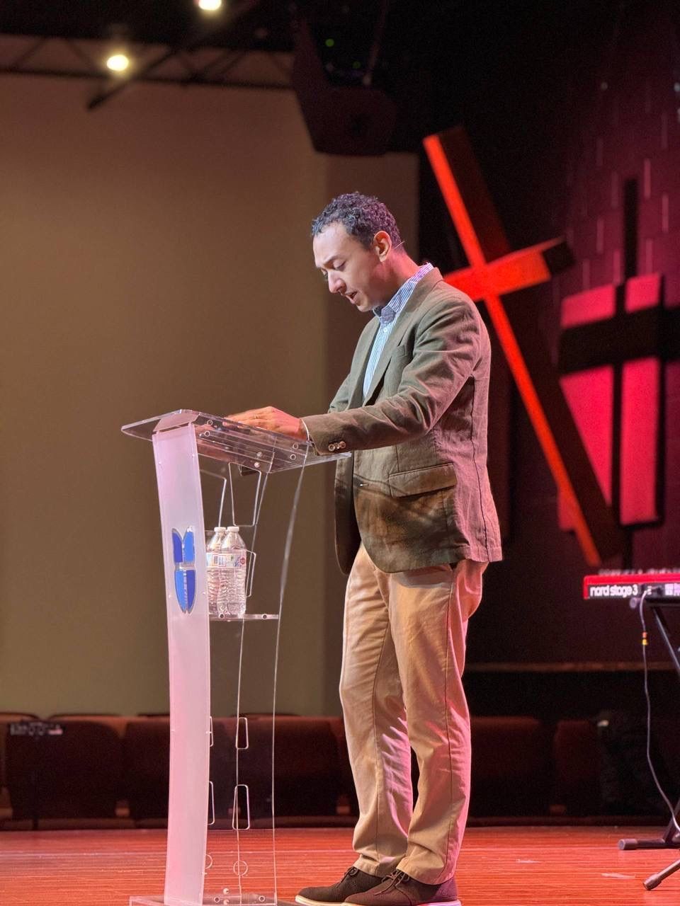 Man speaking from a podium on a stage with a cross backdrop. He is wearing a jacket and khakis.