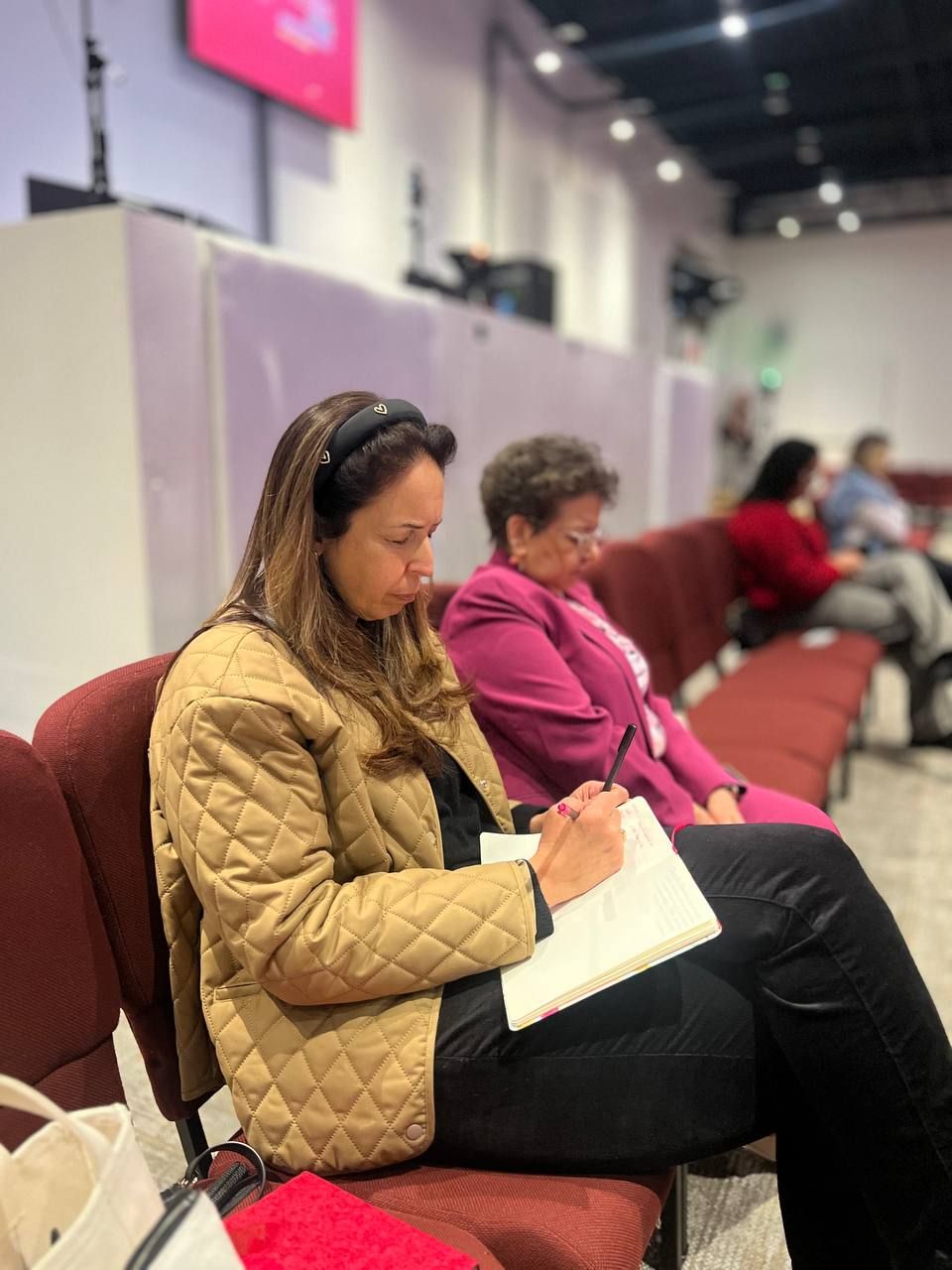 Woman in tan jacket, writing in a notebook in a church pew. Another woman and other people seated nearby.