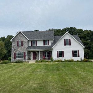 A large white house with a gray roof and red shutters is sitting on top of a lush green field.