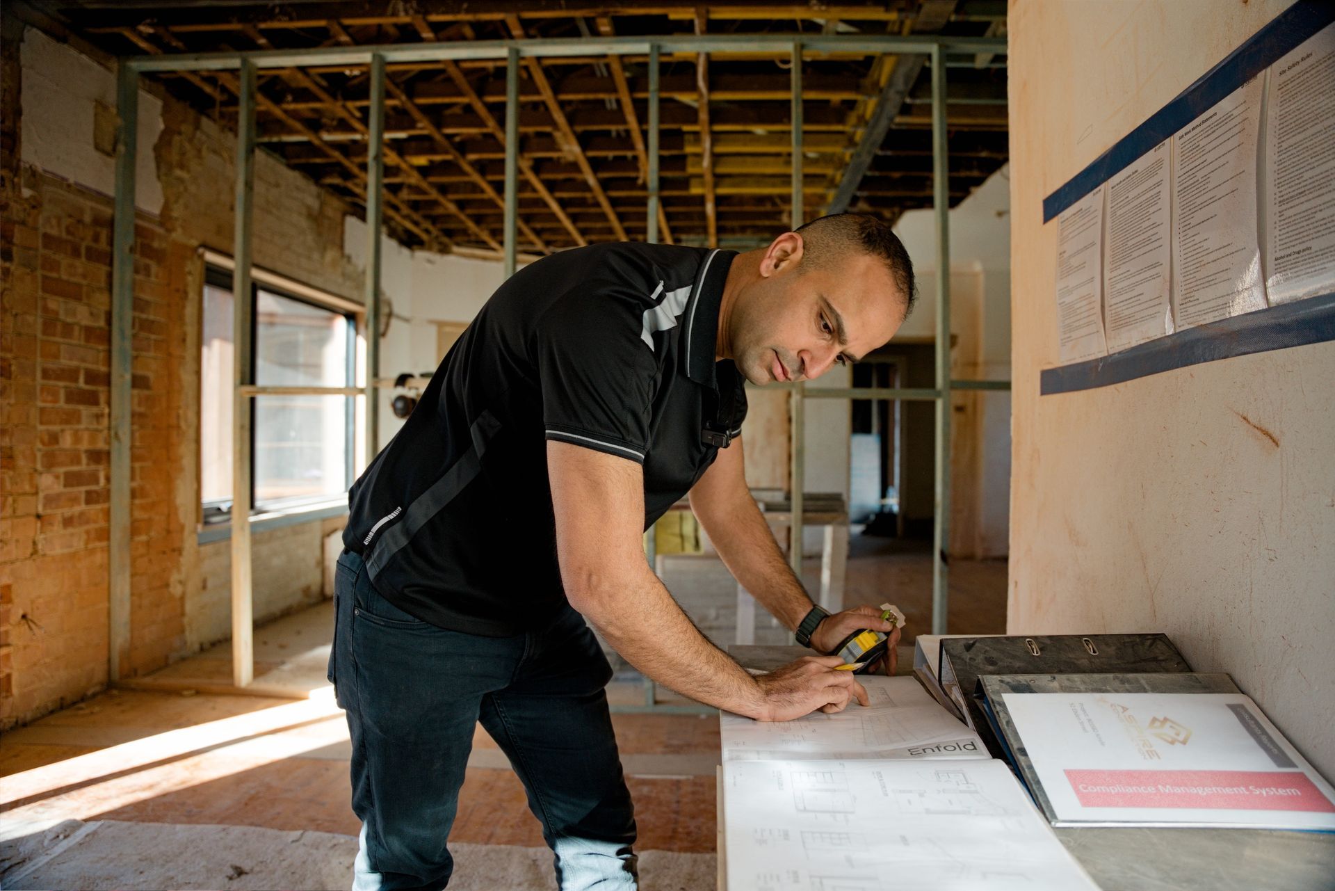 Man measuring and marking wood in an unfinished room with exposed framing and brick walls