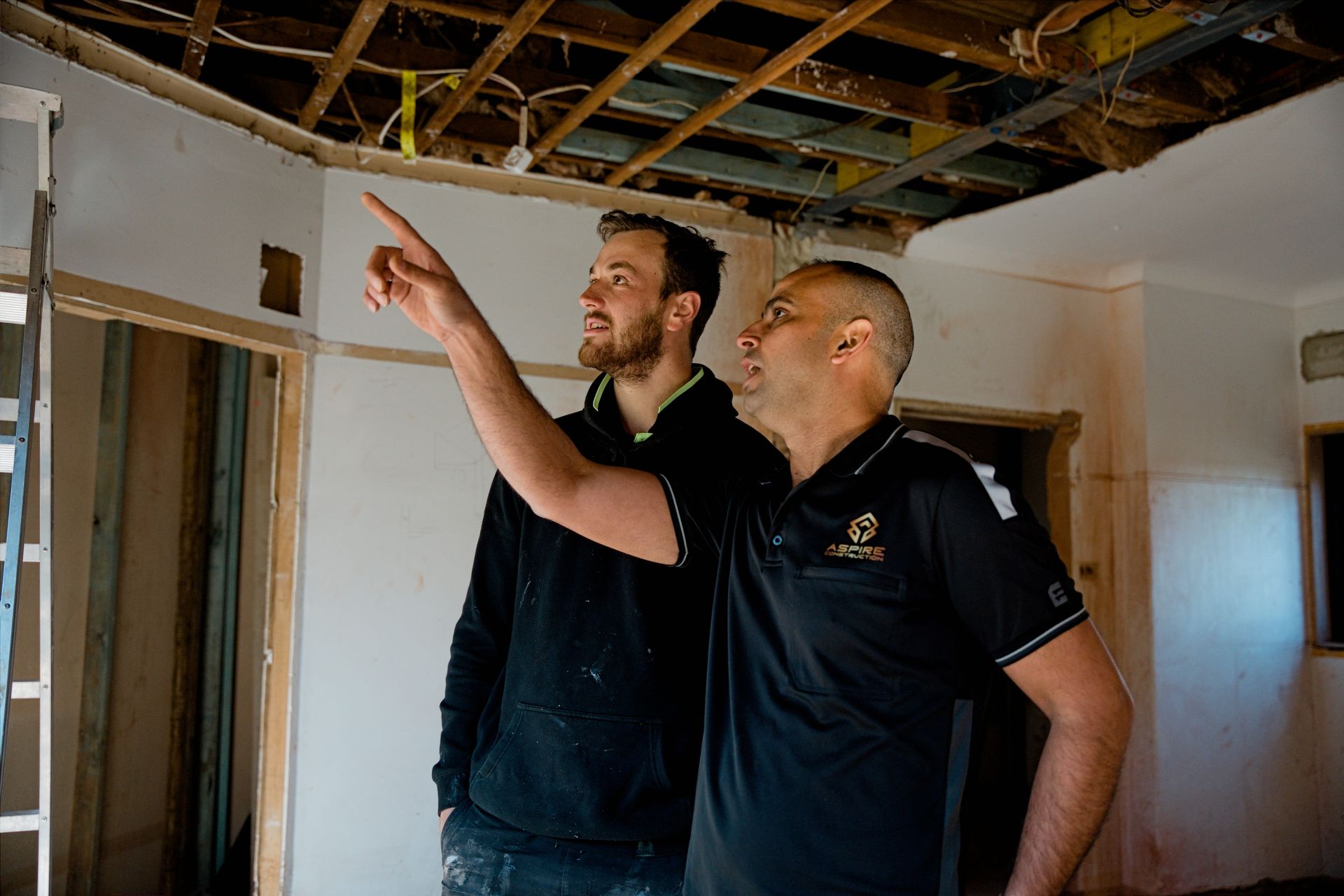 Two men in black shirts inspecting exposed ceiling framing in a room under renovation