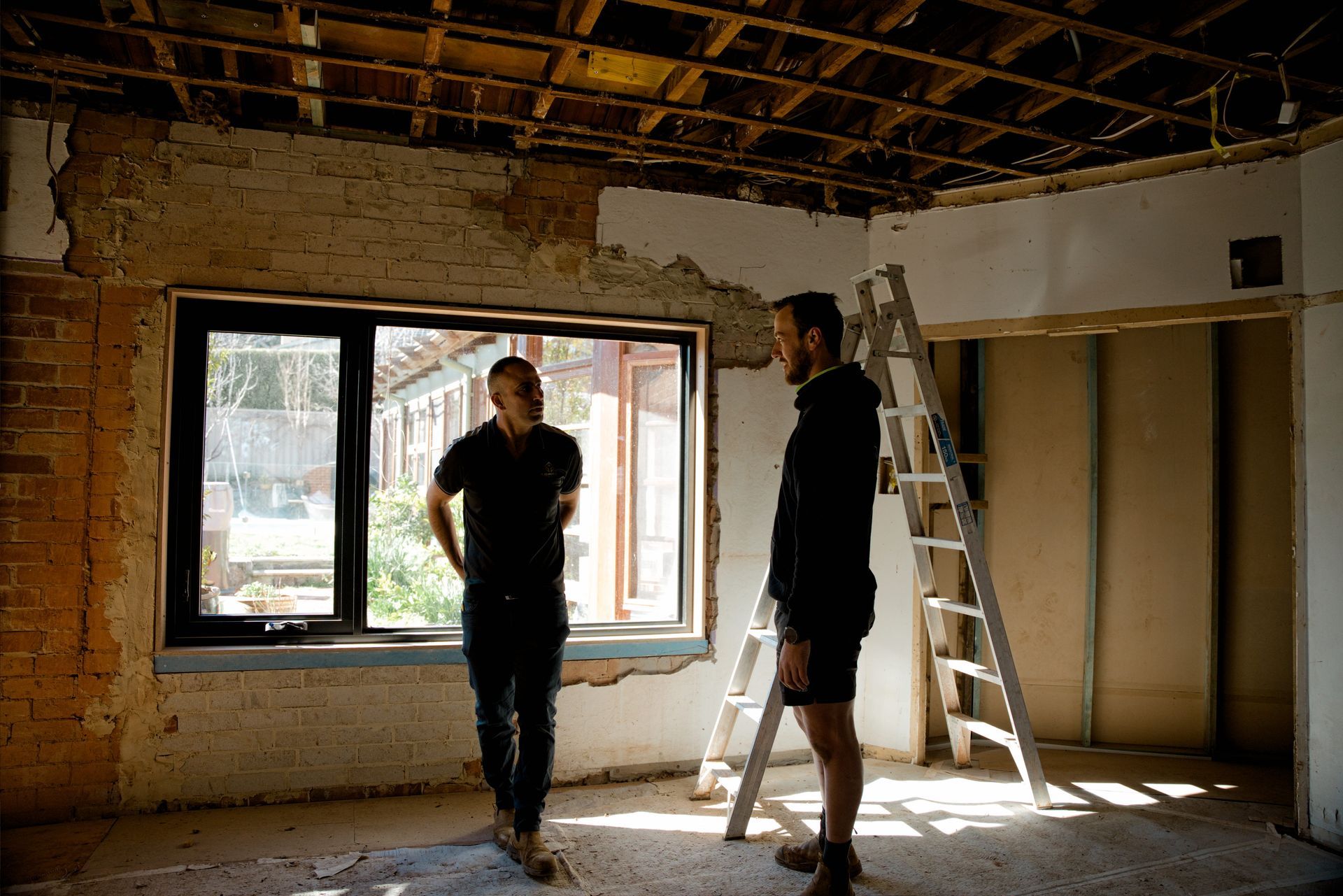 Two people standing in a dimly lit room under renovation, with exposed ceiling beams and a ladder by a window