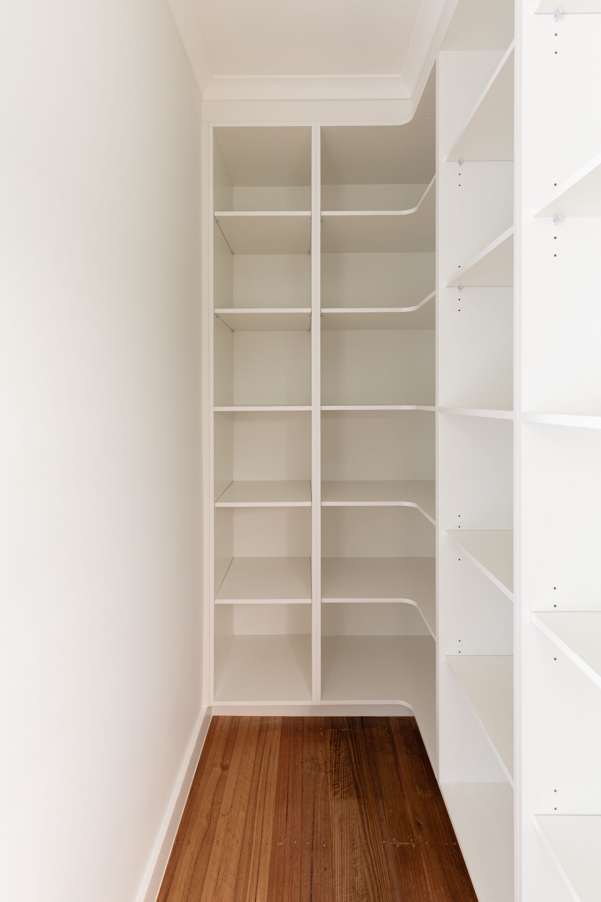 Narrow walk-in closet with white shelves and drawers on both sides, wood floor, and white walls.