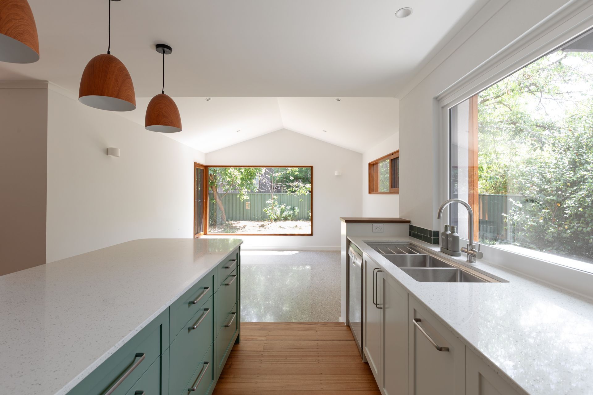 Bright modern kitchen with white counters, green cabinets, and large windows overlooking greenery.