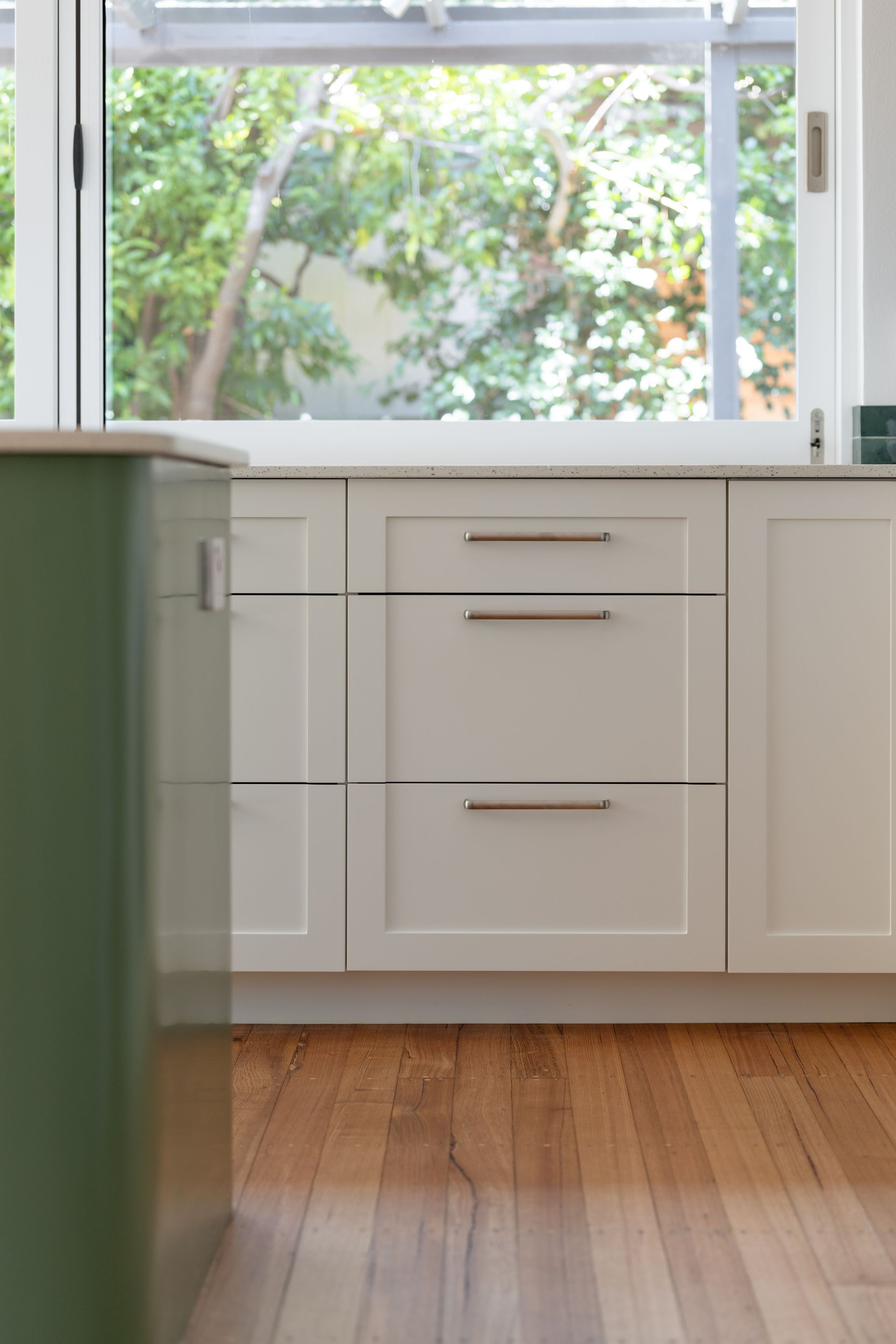 Bright kitchen with white cabinets, wood floor, and a large window overlooking greenery.