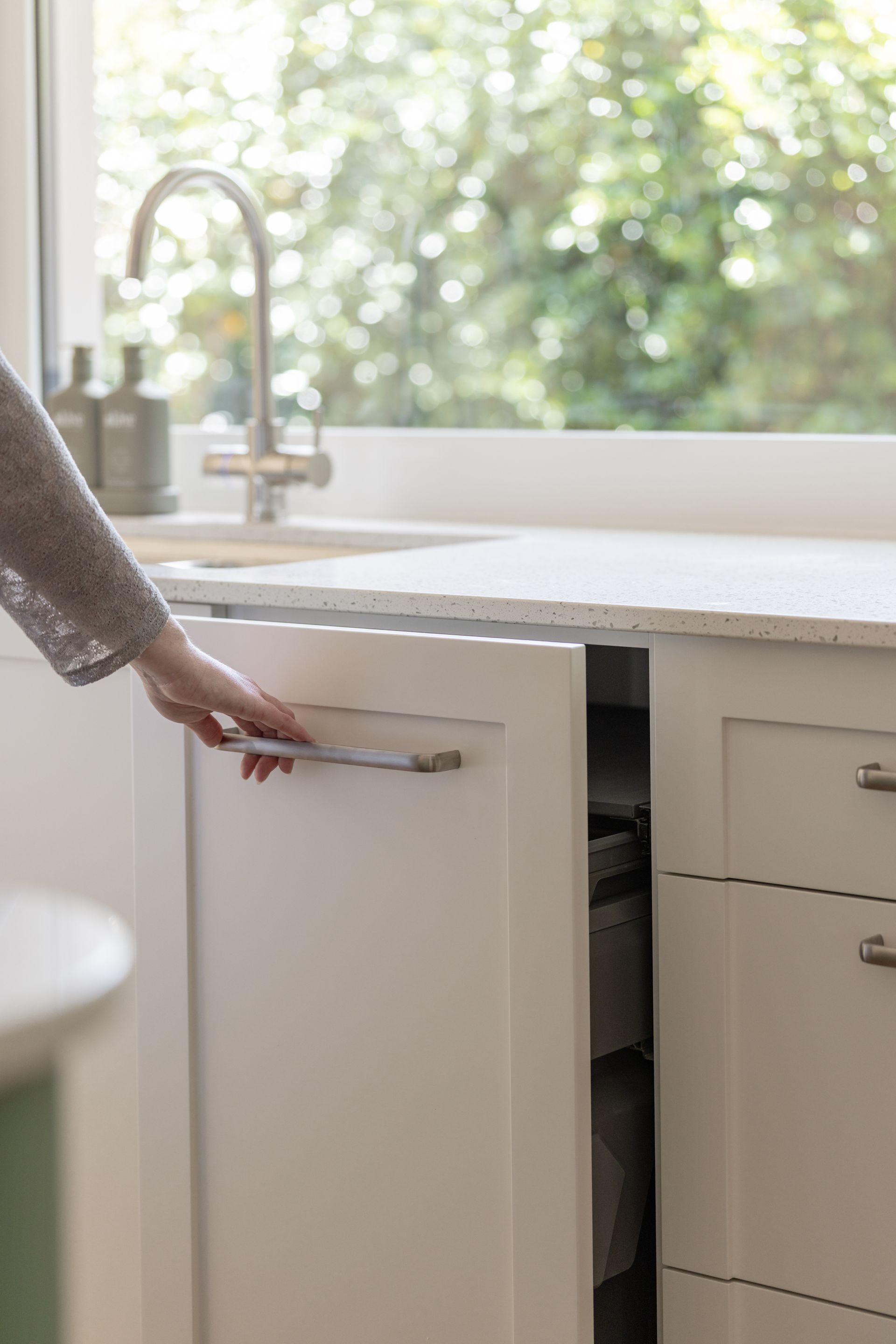 Hand opening a white kitchen cabinet under a sink in a bright, modern kitchen.