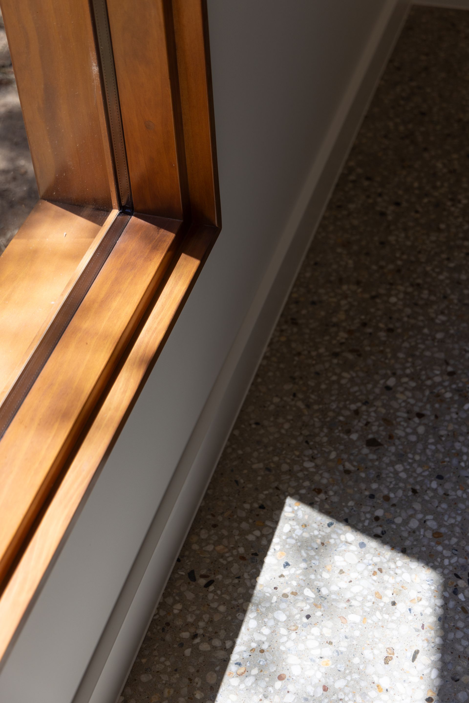 Sunlight on a speckled floor beside a wooden window frame and white wall.