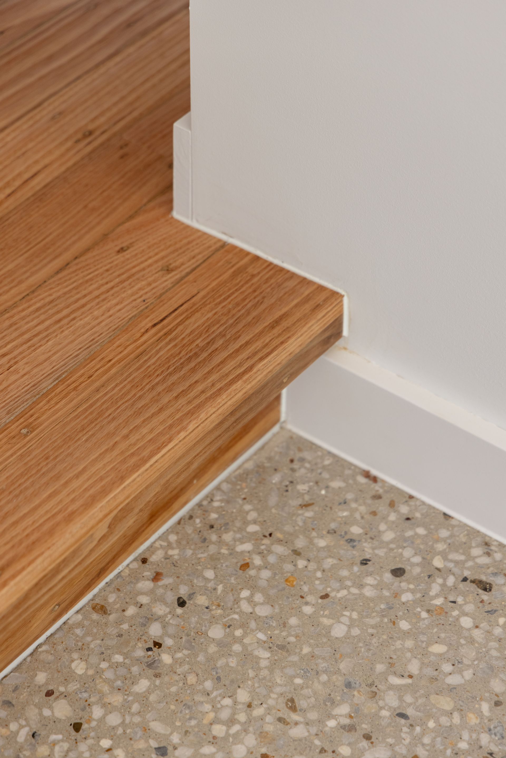 Wooden stair edge beside a white wall over a speckled terrazzo floor