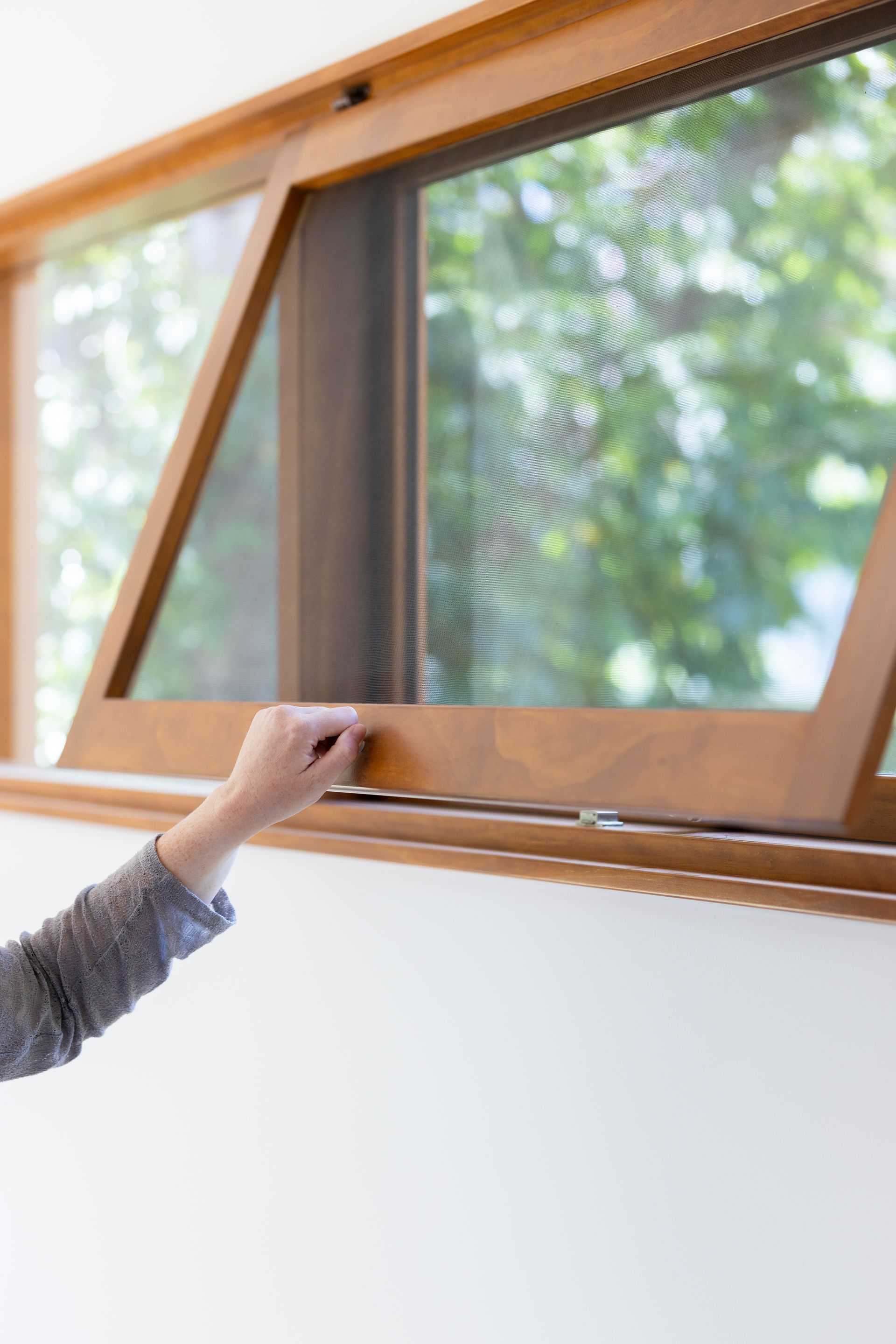 Hand opening a wooden awning window, with green trees visible outside