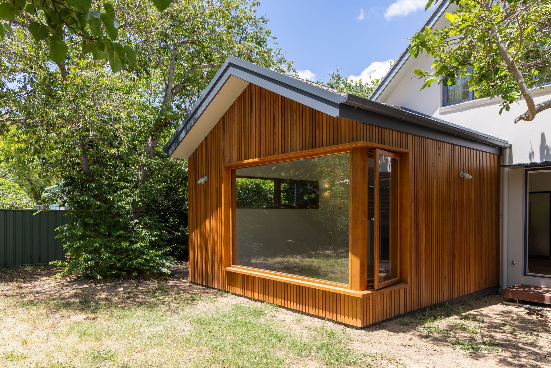 Modern wooden garden room with large corner windows beside a house, surrounded by trees and grass