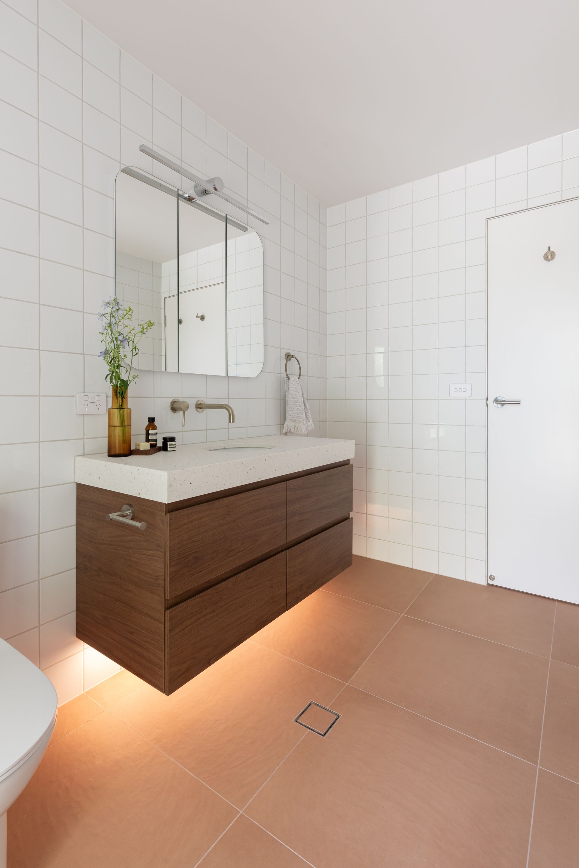 Modern white-tiled bathroom with wood vanity, mirror, sink, shower, and tan floor tiles