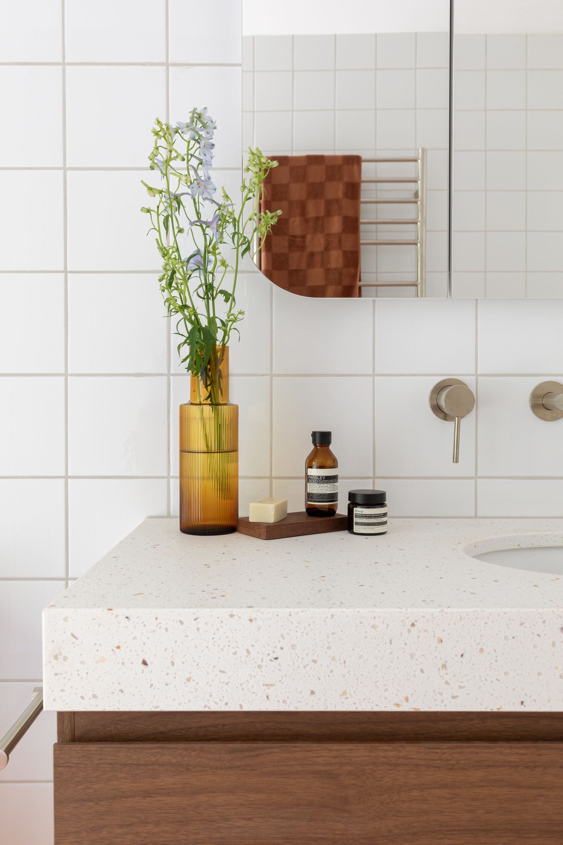 Minimal bathroom vanity with amber vase flowers, soap bottles, and towels against white tiled wall