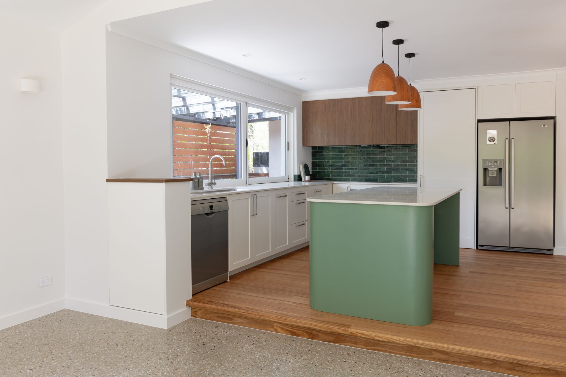 Modern white kitchen with green island, wooden stools, stainless fridge, and wood flooring