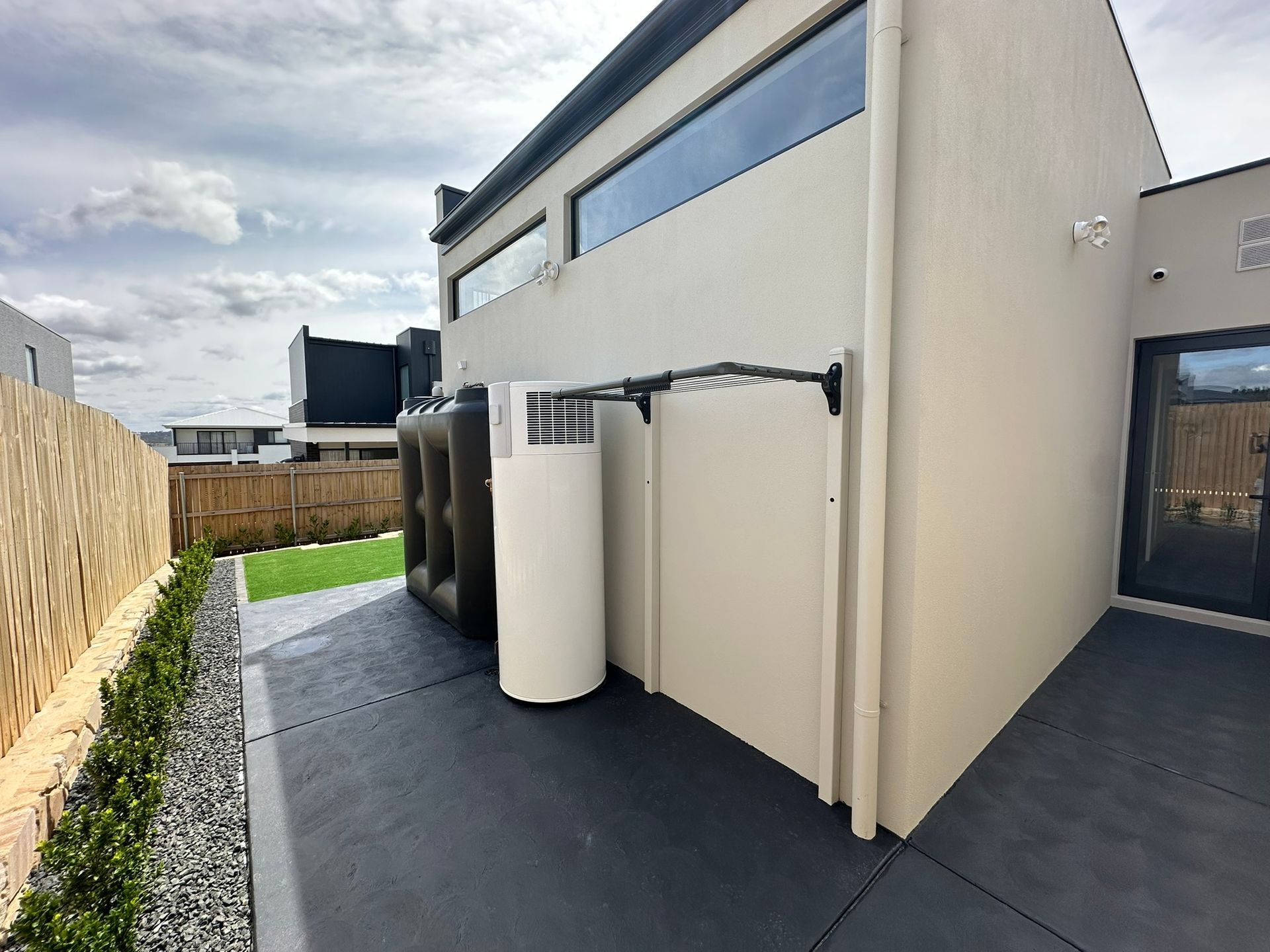 Narrow side yard with cream exterior wall, black deck, white utility tanks, and wooden fence under cloudy sky