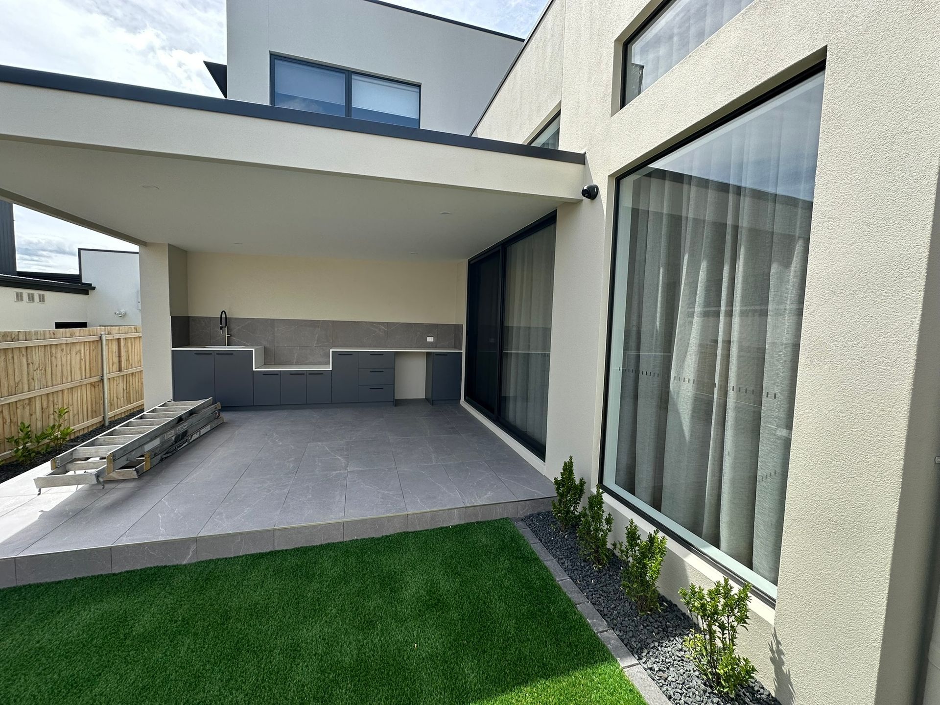 Modern patio with covered seating area, gray tiles, sliding glass doors, and green lawn in front of a house