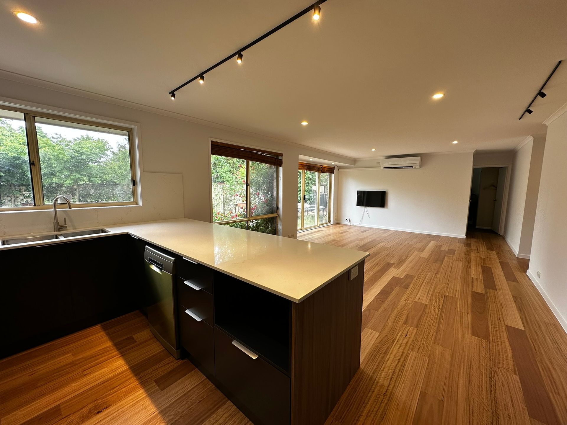 Modern open-plan kitchen and living room with wood floors, dark cabinets, large windows, and recessed lighting