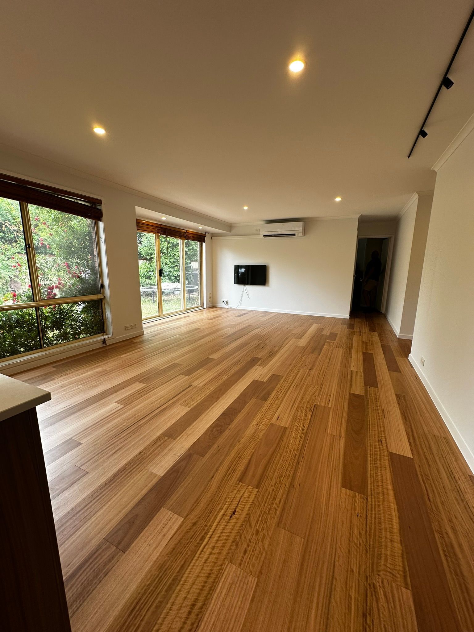 Empty modern living room with wood floor, recessed lights, wall-mounted TV, and large windows overlooking greenery