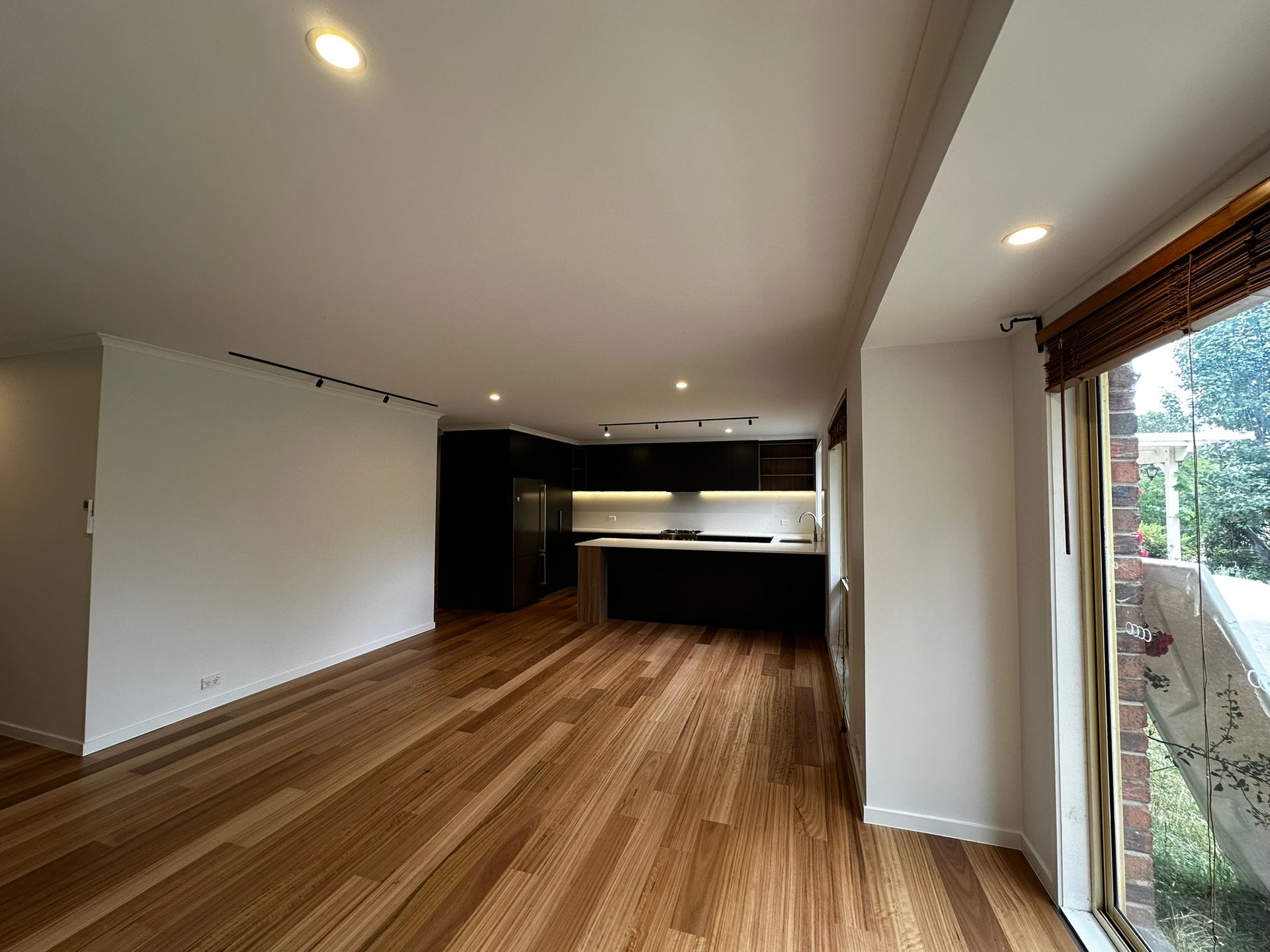 Empty modern living room with wood floors, recessed lights, and kitchen area at the back.