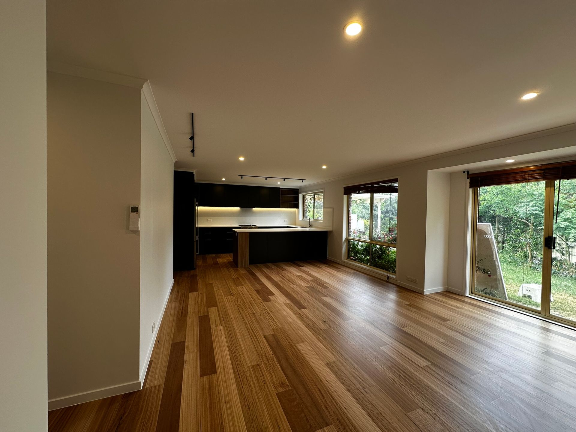 Empty modern living room with wood floors, kitchen at back, and large windows overlooking greenery