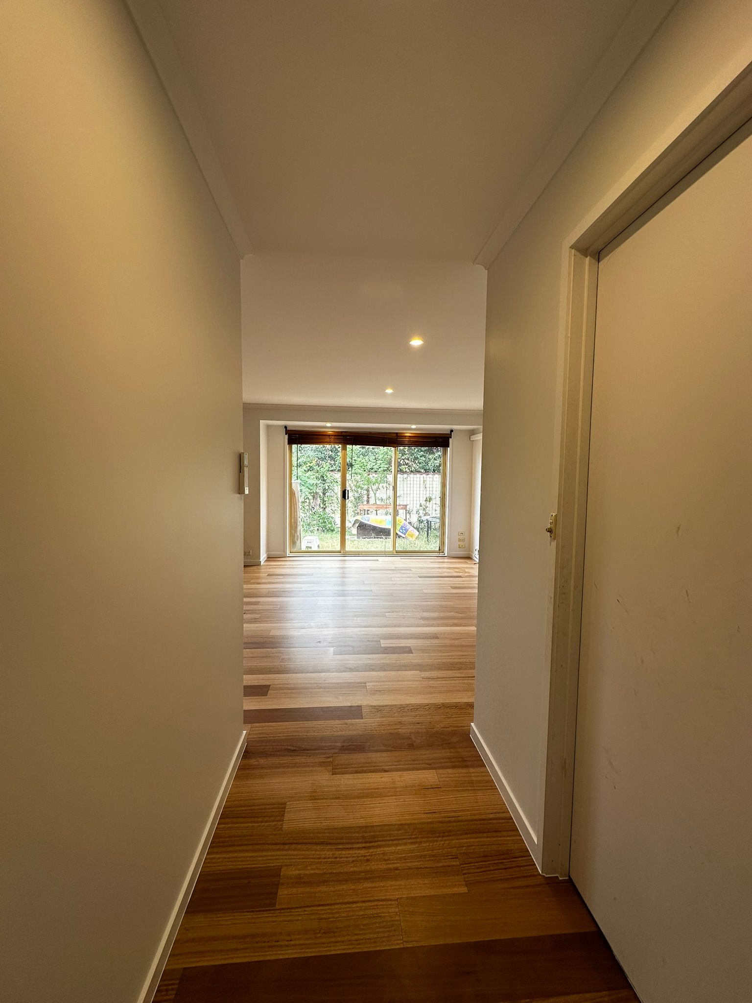 Narrow hallway with white walls and wood floor leading to a bright living room with glass doors to greenery
