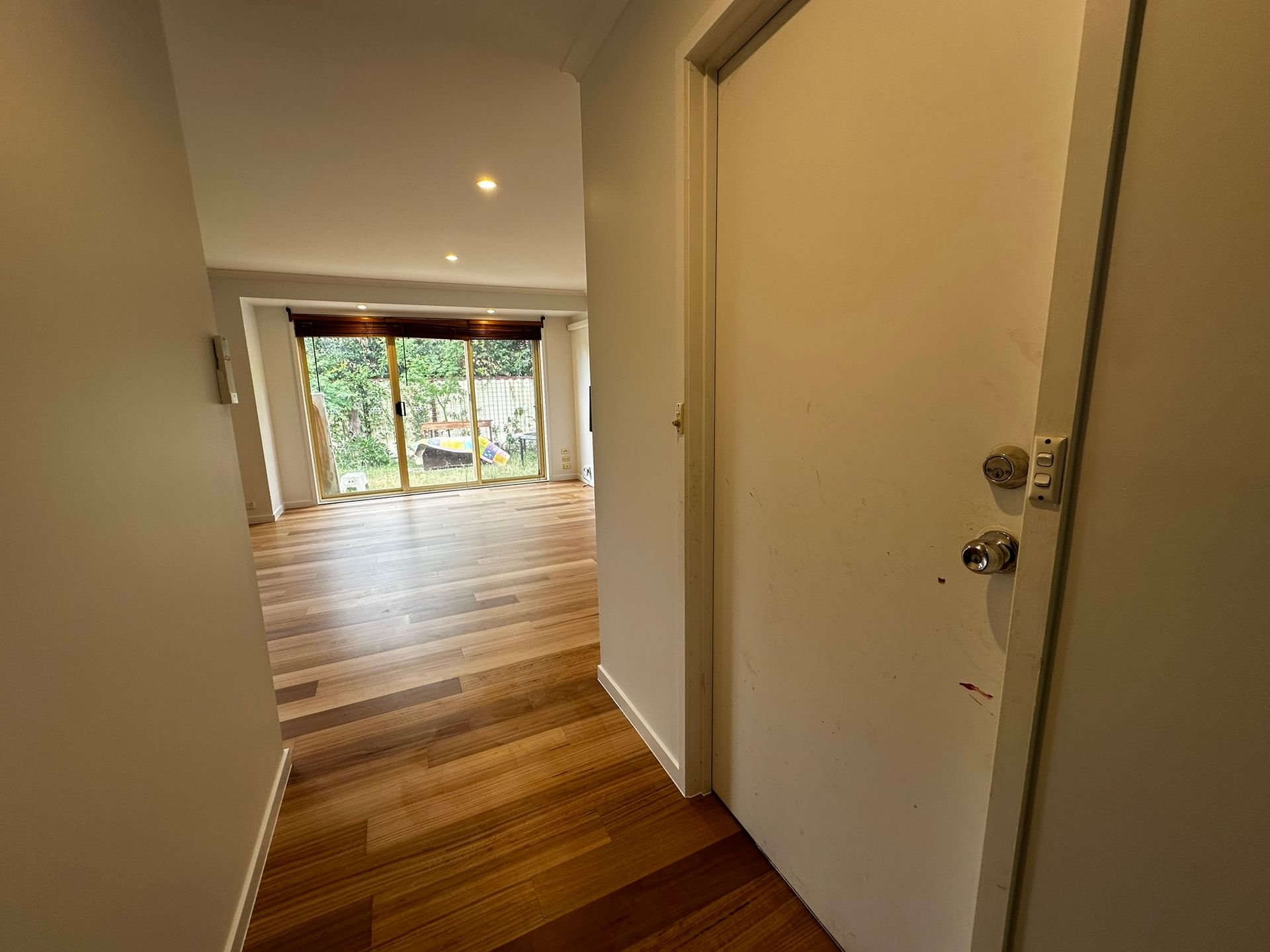Narrow hallway with white walls and wood floor leading to a bright room with sliding glass doors.