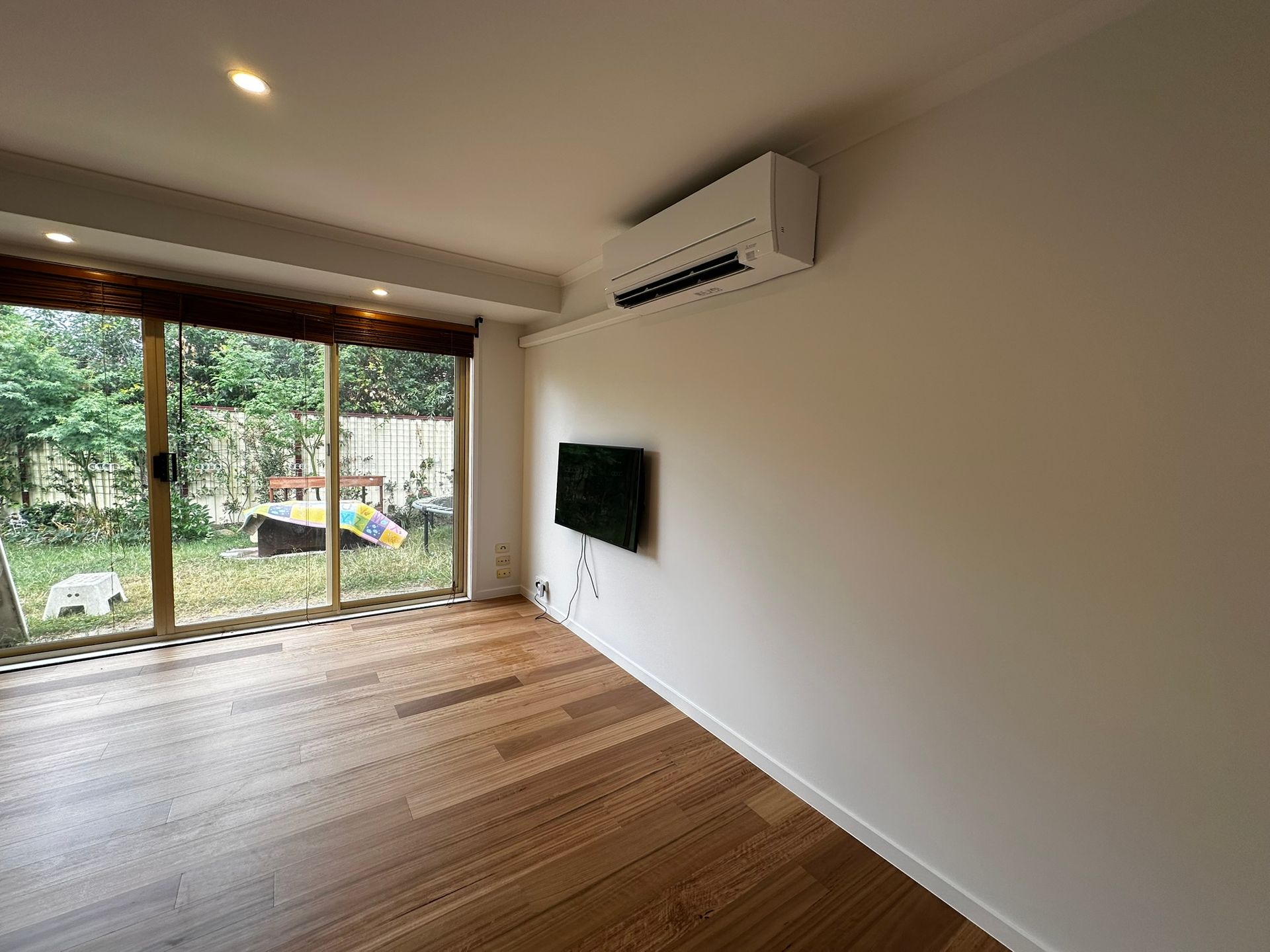 Empty modern living room with wood floors, wall-mounted TV, and large sliding doors to a green yard.