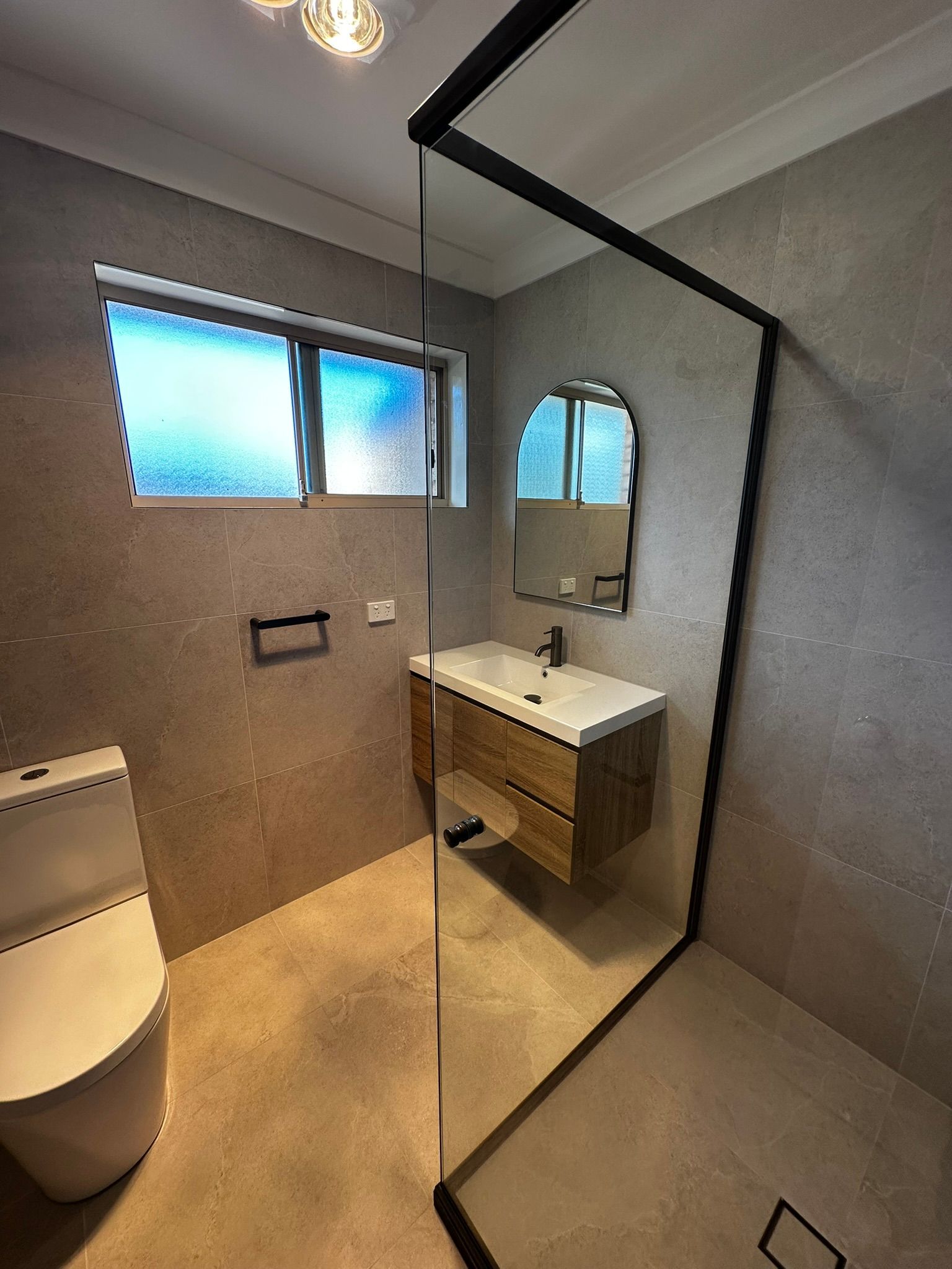 Modern bathroom with toilet, sink, mirror, shower glass, and frosted window.
