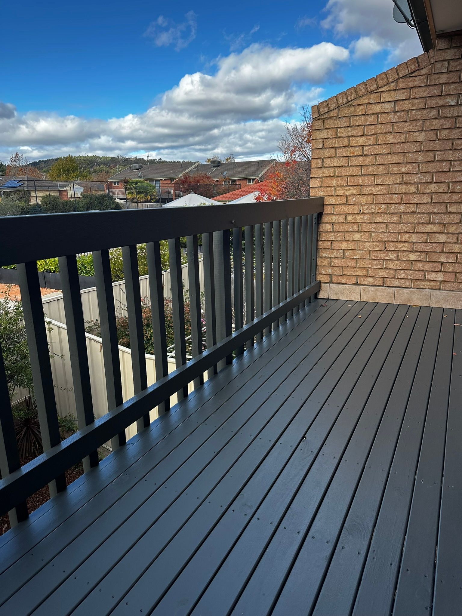 Balcony with dark railing and gray floor beside tan brick wall, overlooking trees and cloudy sky