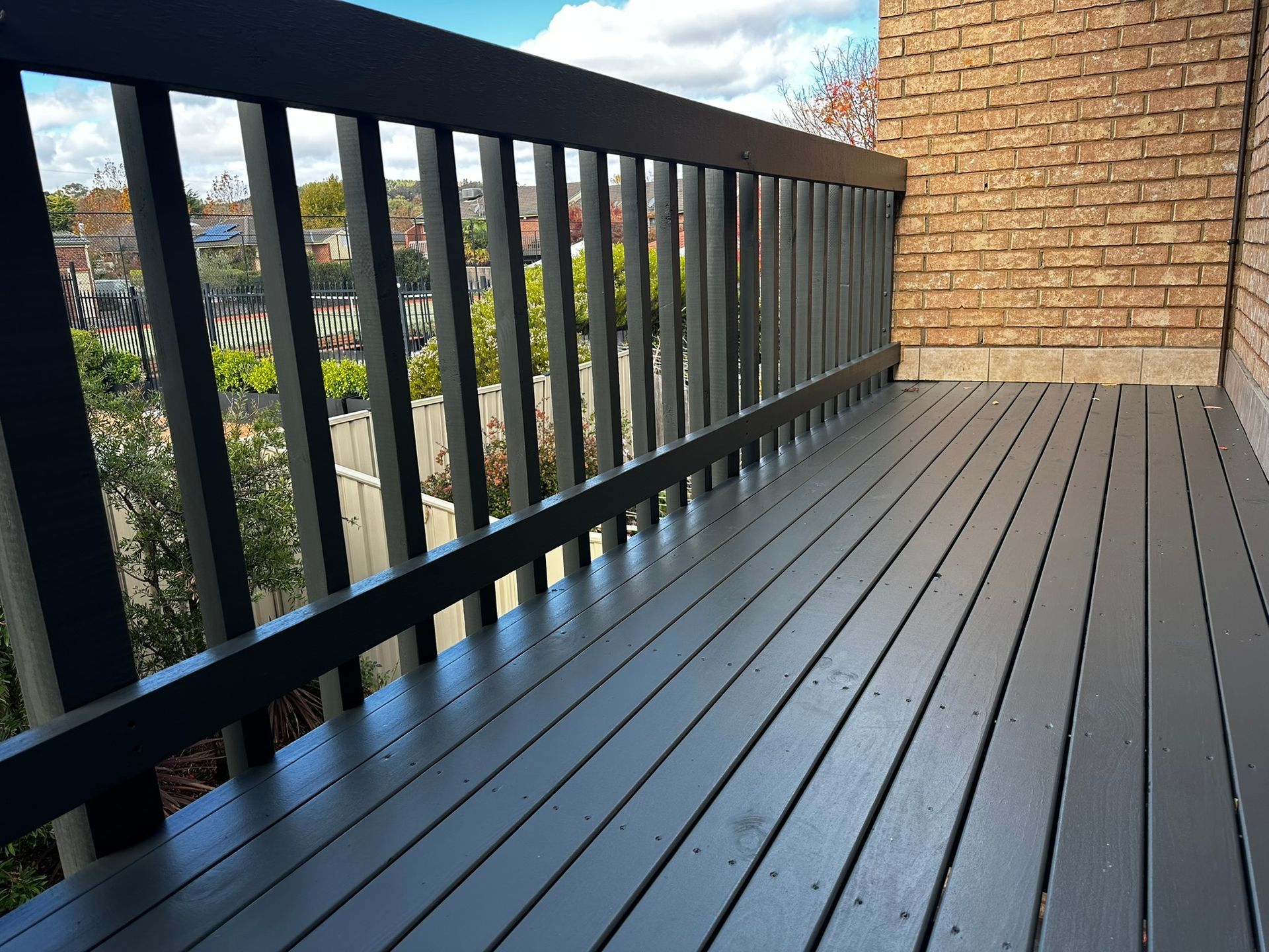 Balcony with dark railing and gray deck beside a brick wall, overlooking trees and buildings