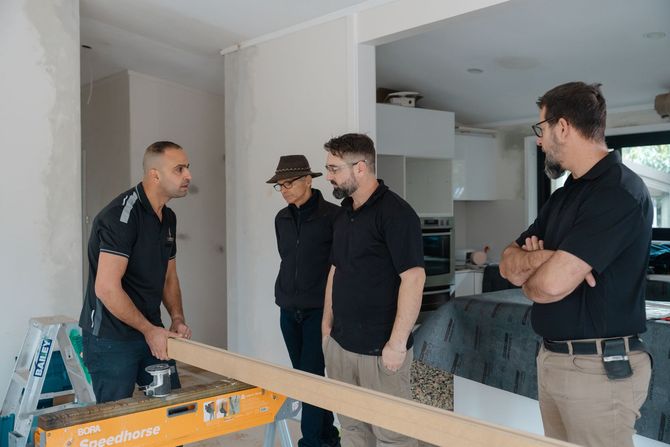 Four men stand in a partially renovated room, talking near tools and unfinished walls.