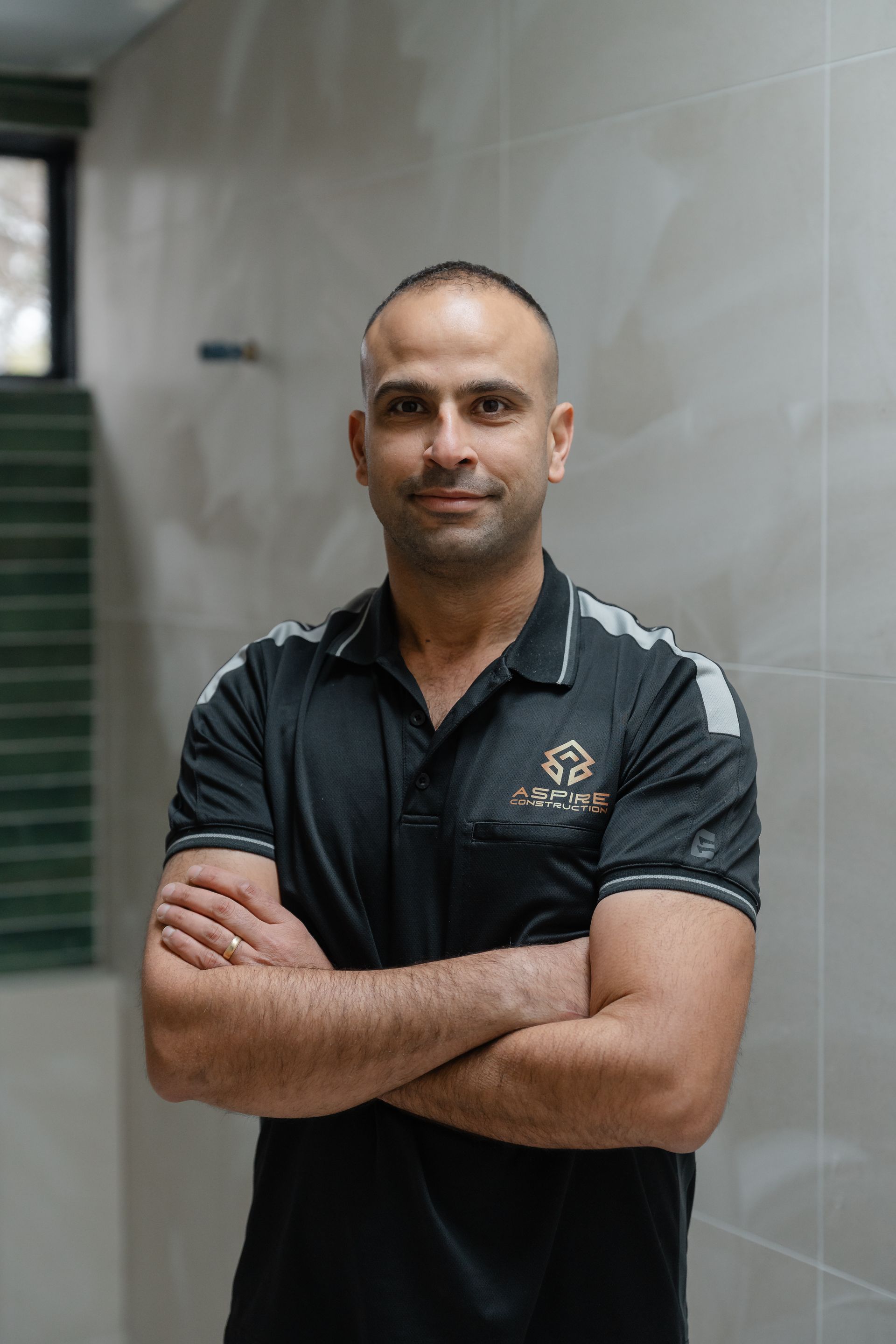 Man standing with arms crossed in a black polo shirt against a tiled indoor wall.