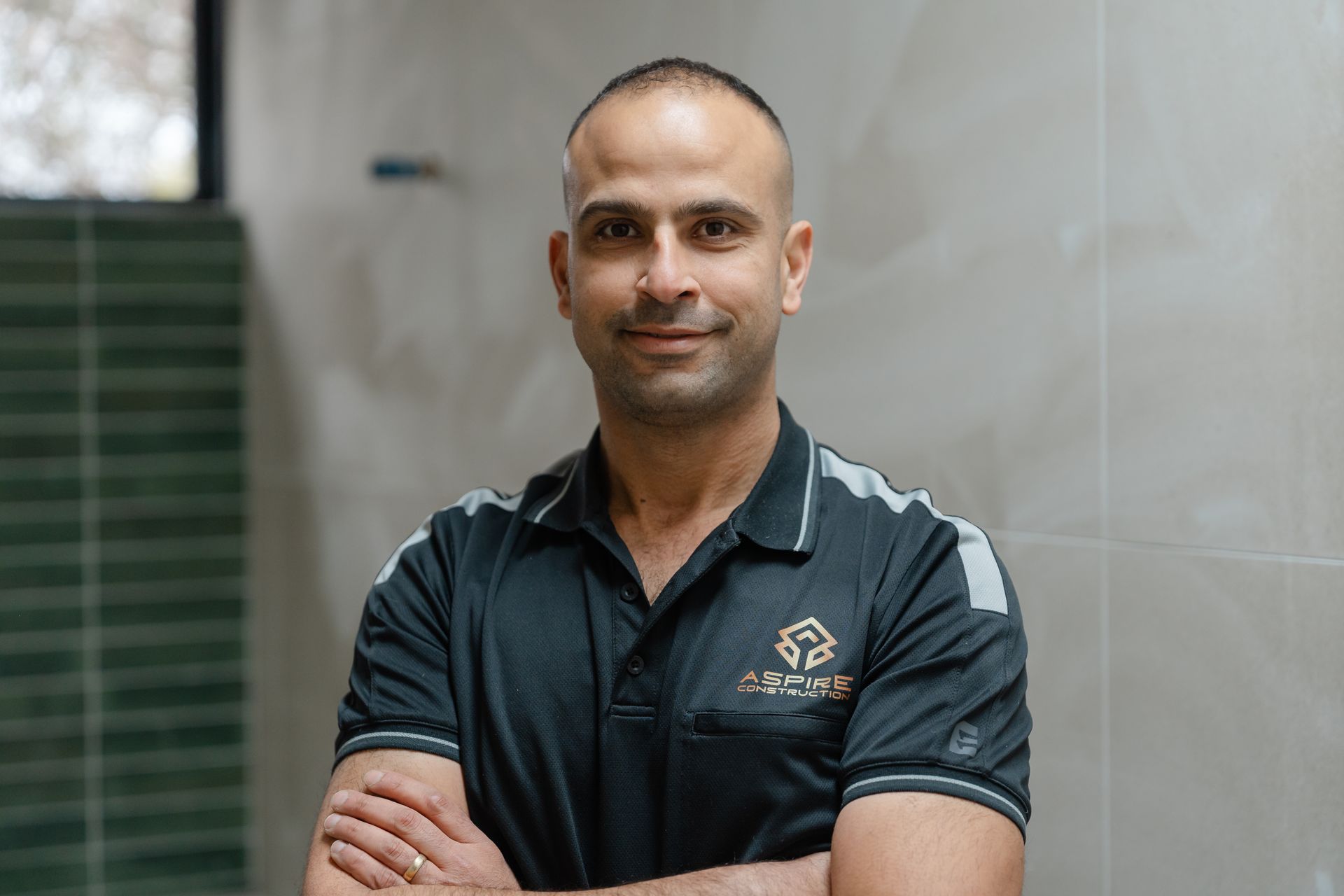 Man standing with arms crossed in a dark polo shirt against a tiled indoor wall.
