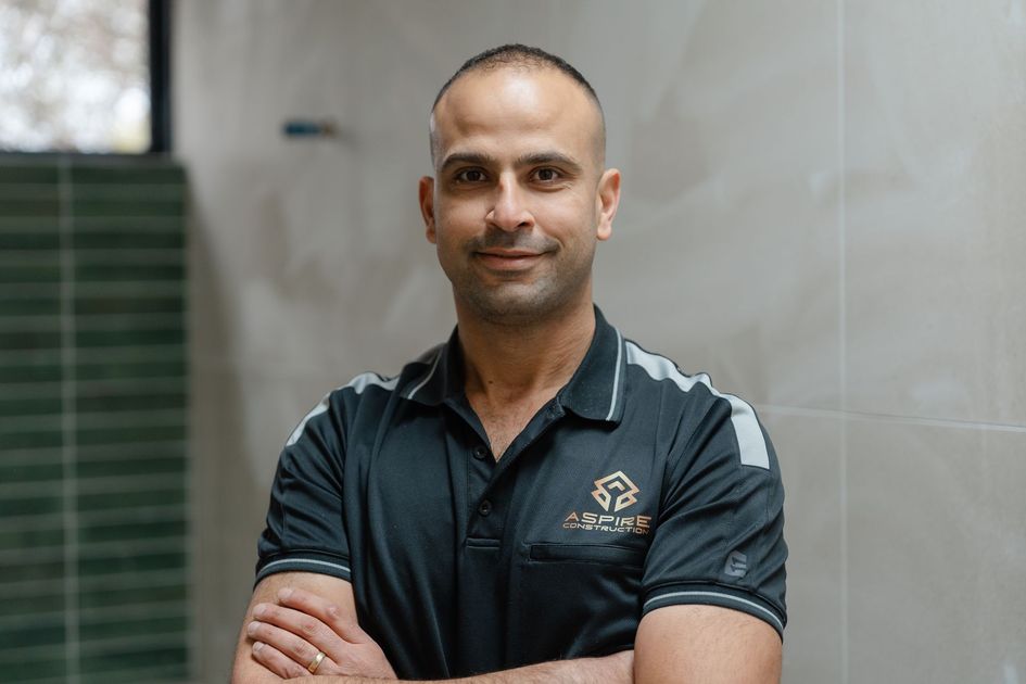 Man standing with arms crossed in a dark polo shirt against a tiled indoor wall.