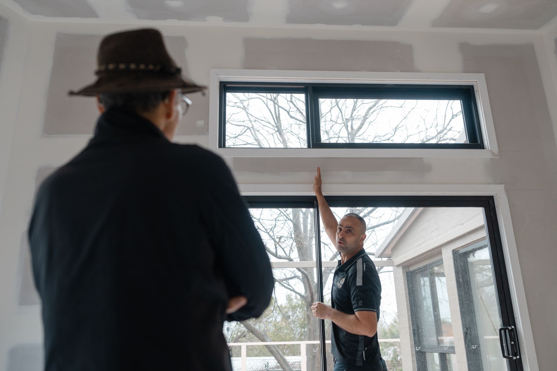 Two people discussing a window installation in a bright room, one pointing upward near the frame.
