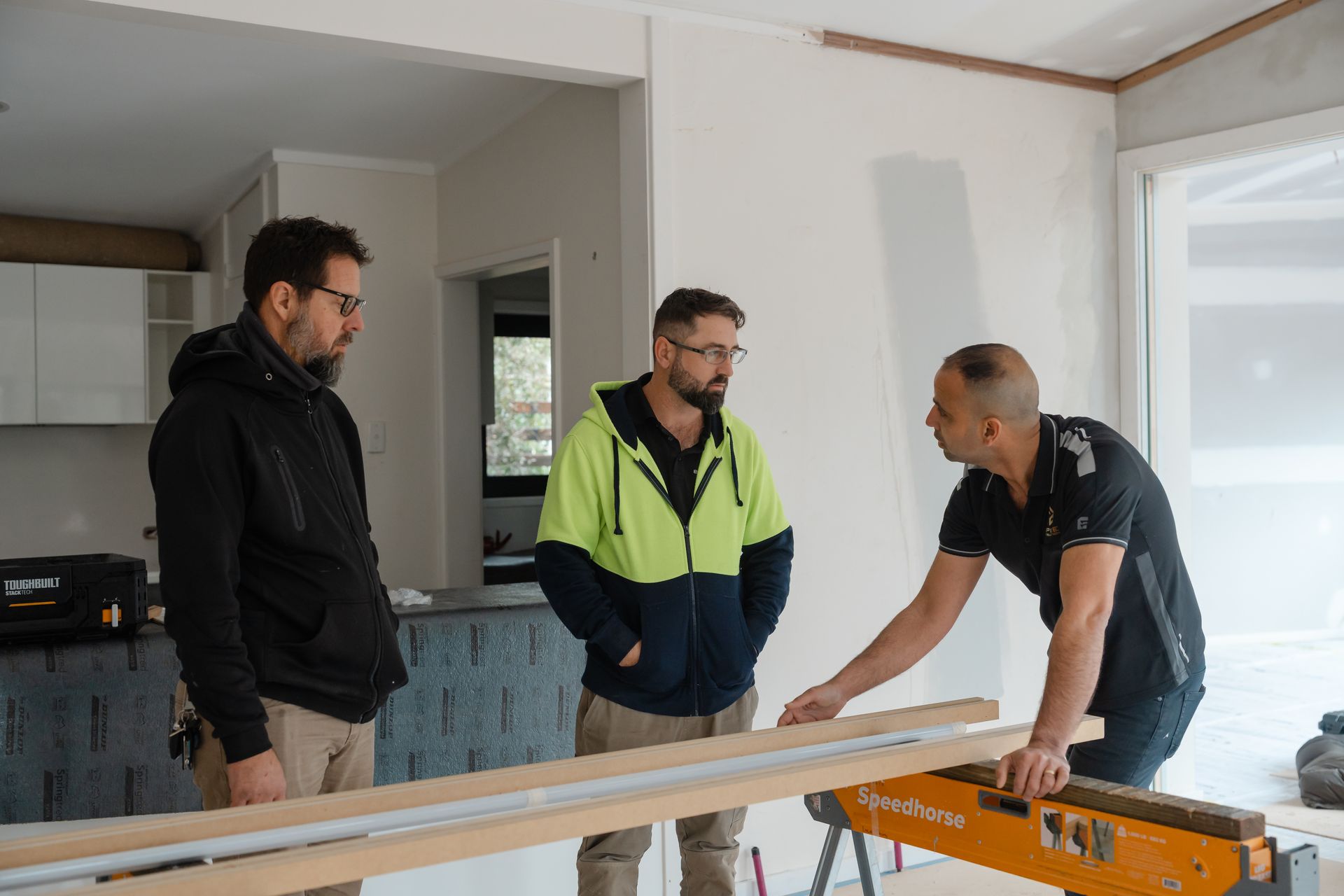 Three men measuring and discussing a wooden plank at a construction site indoors
