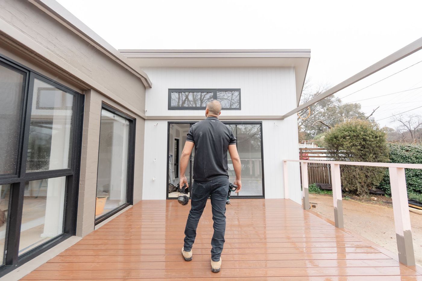 Person walking on a wooden deck beside a modern house with large windows.