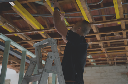 Person on a ladder working overhead in an unfinished room with exposed wooden ceiling beams.