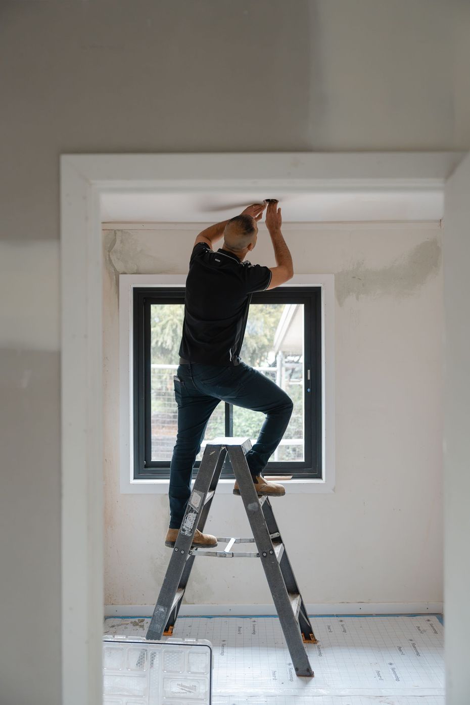 Person on a ladder installing a ceiling fixture in a white room with a window.