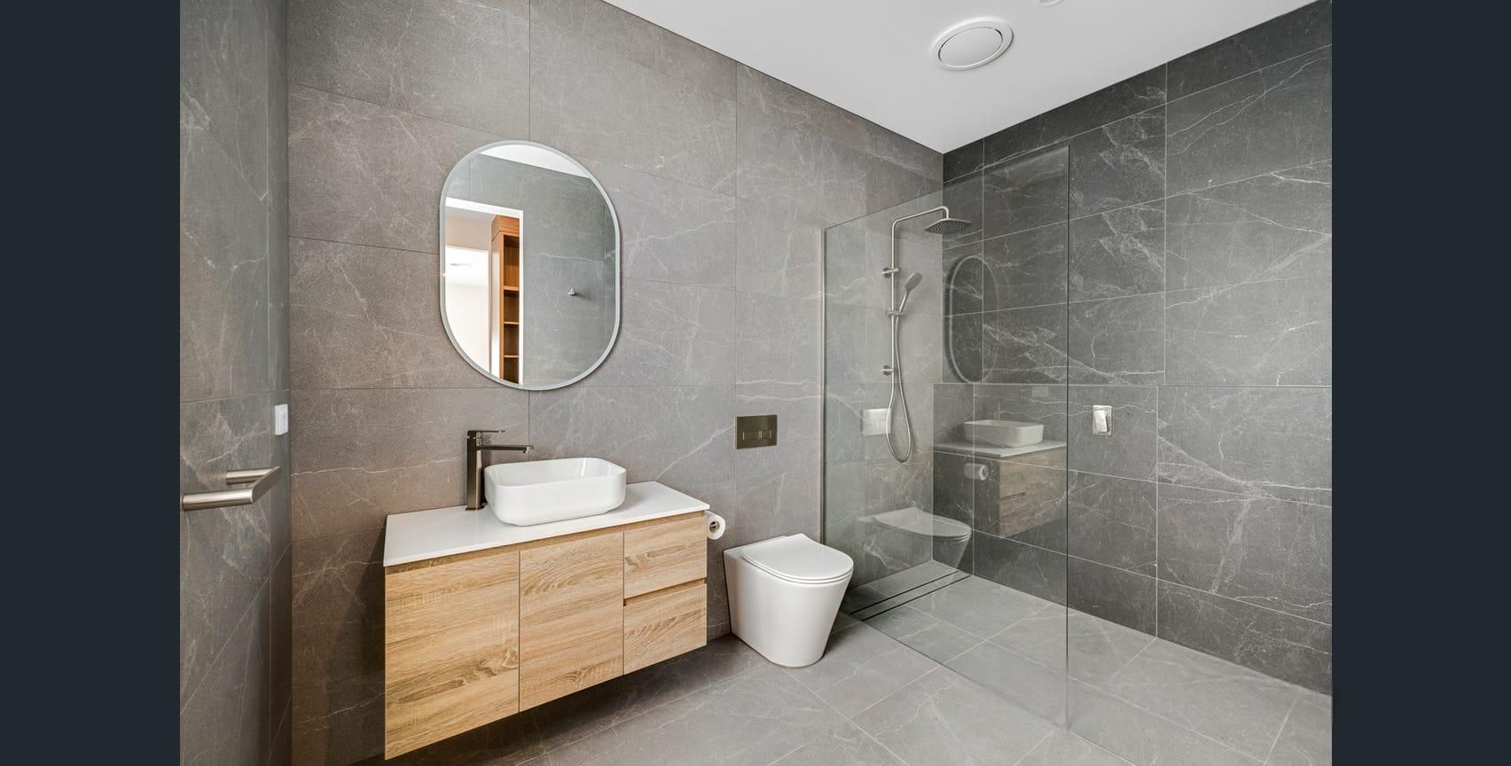 Modern bathroom with wood vanity, round mirror, white toilet, and large gray tiled shower