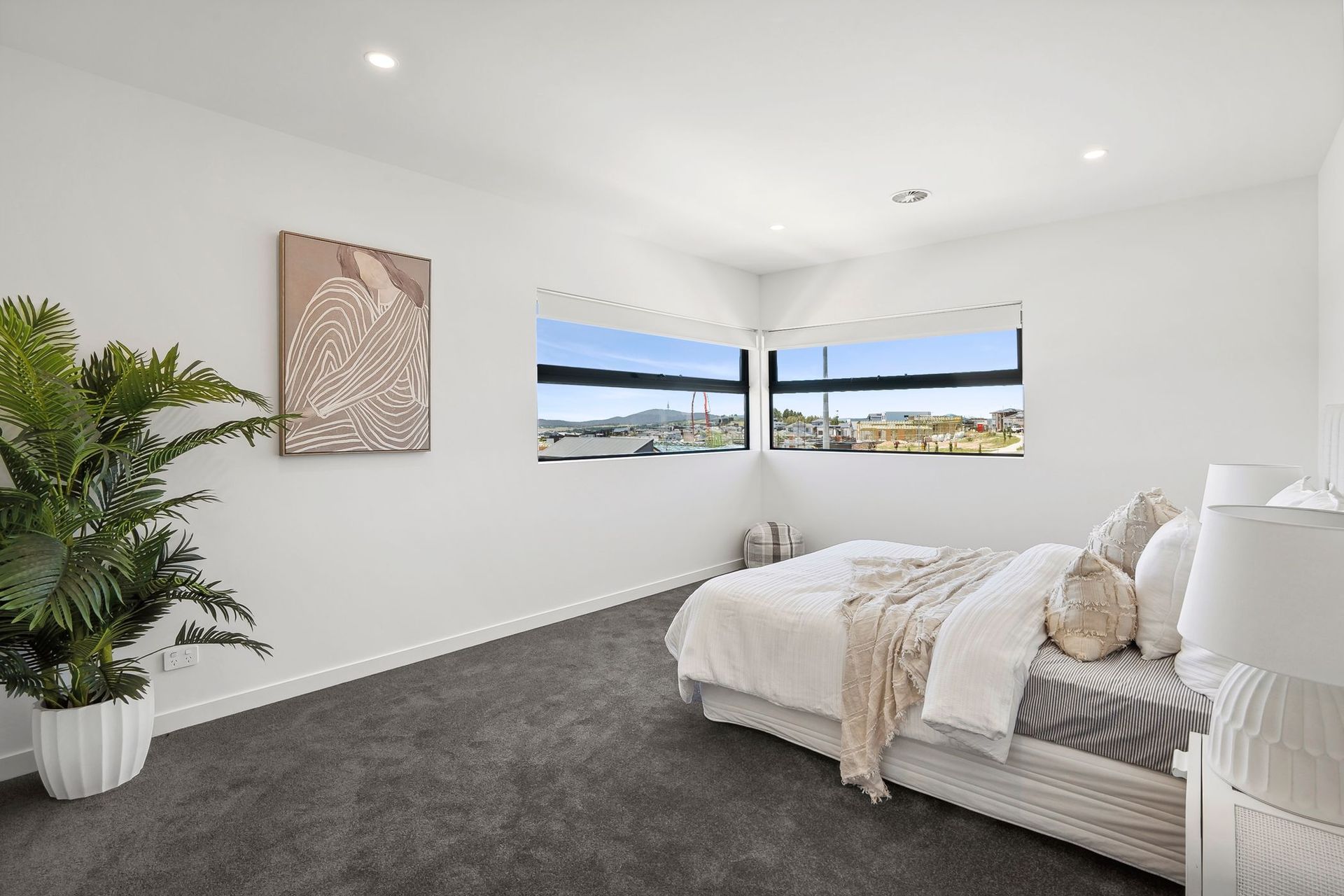 Bright minimalist bedroom with gray carpet, white walls, bed, plant, wall art, and large windows overlooking water.