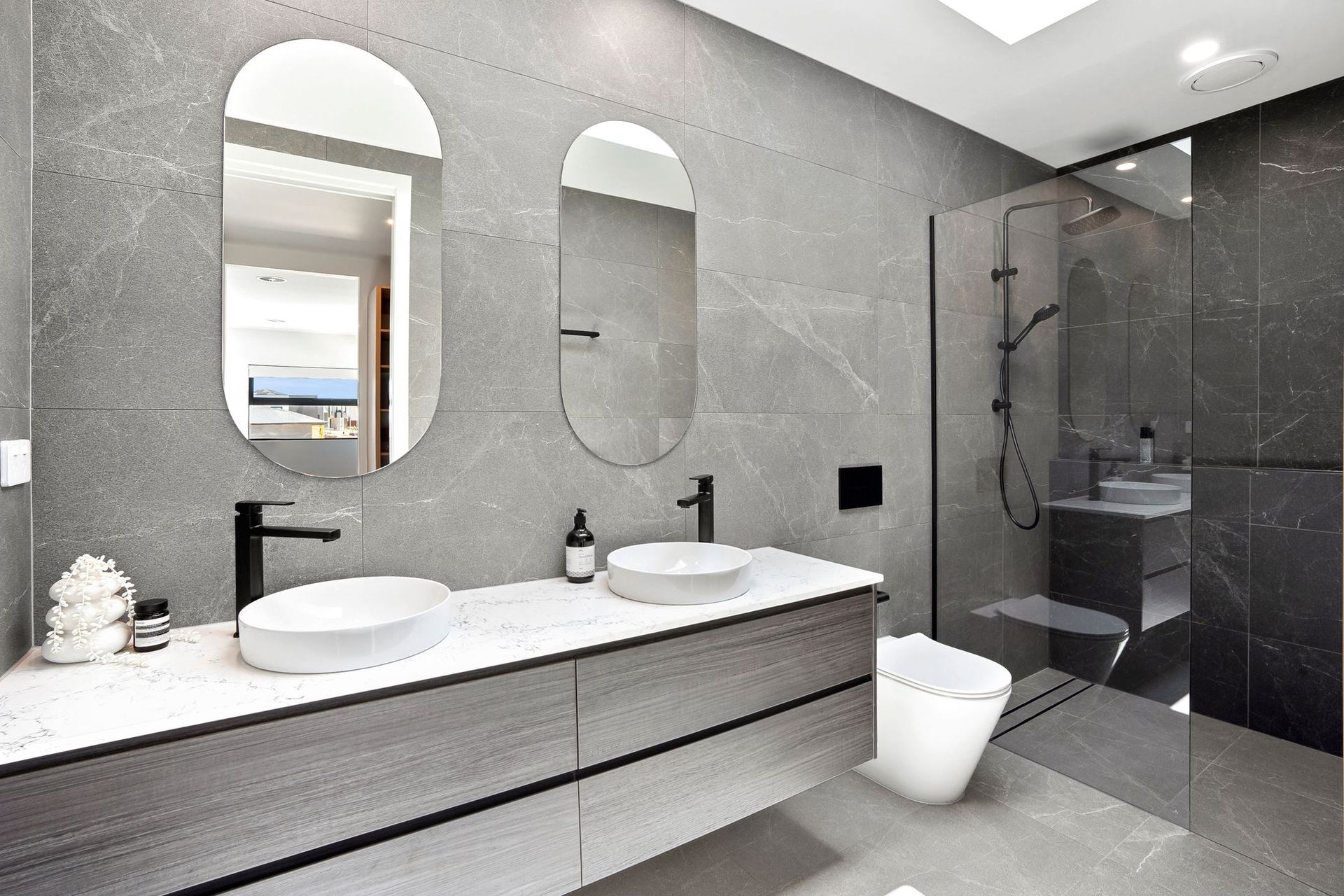 Modern gray bathroom with double sinks, oval mirrors, and a glass-enclosed shower.