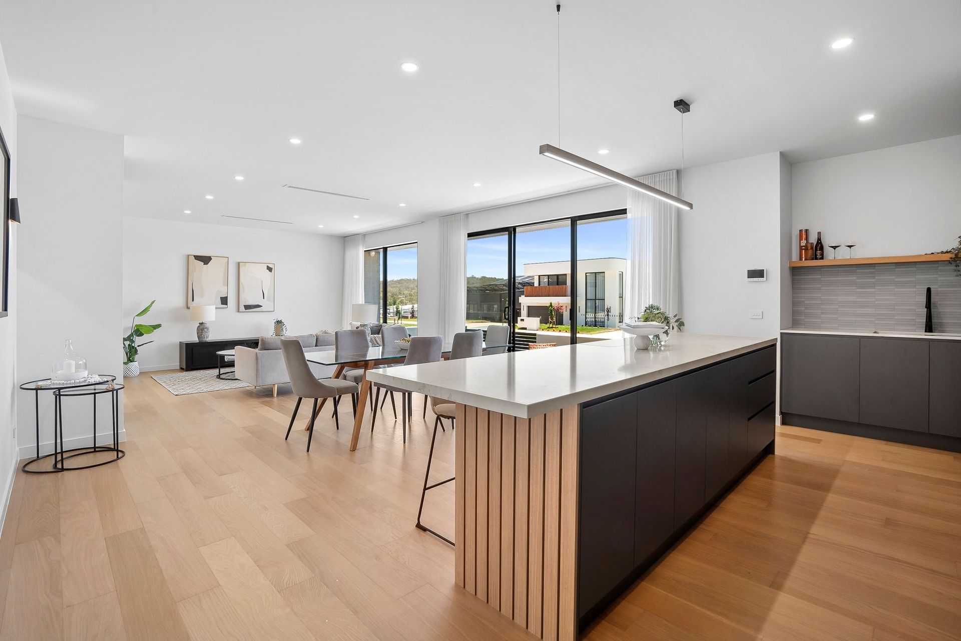 Bright modern open-plan kitchen and dining area with island, white cabinets, wood floors, and large windows.